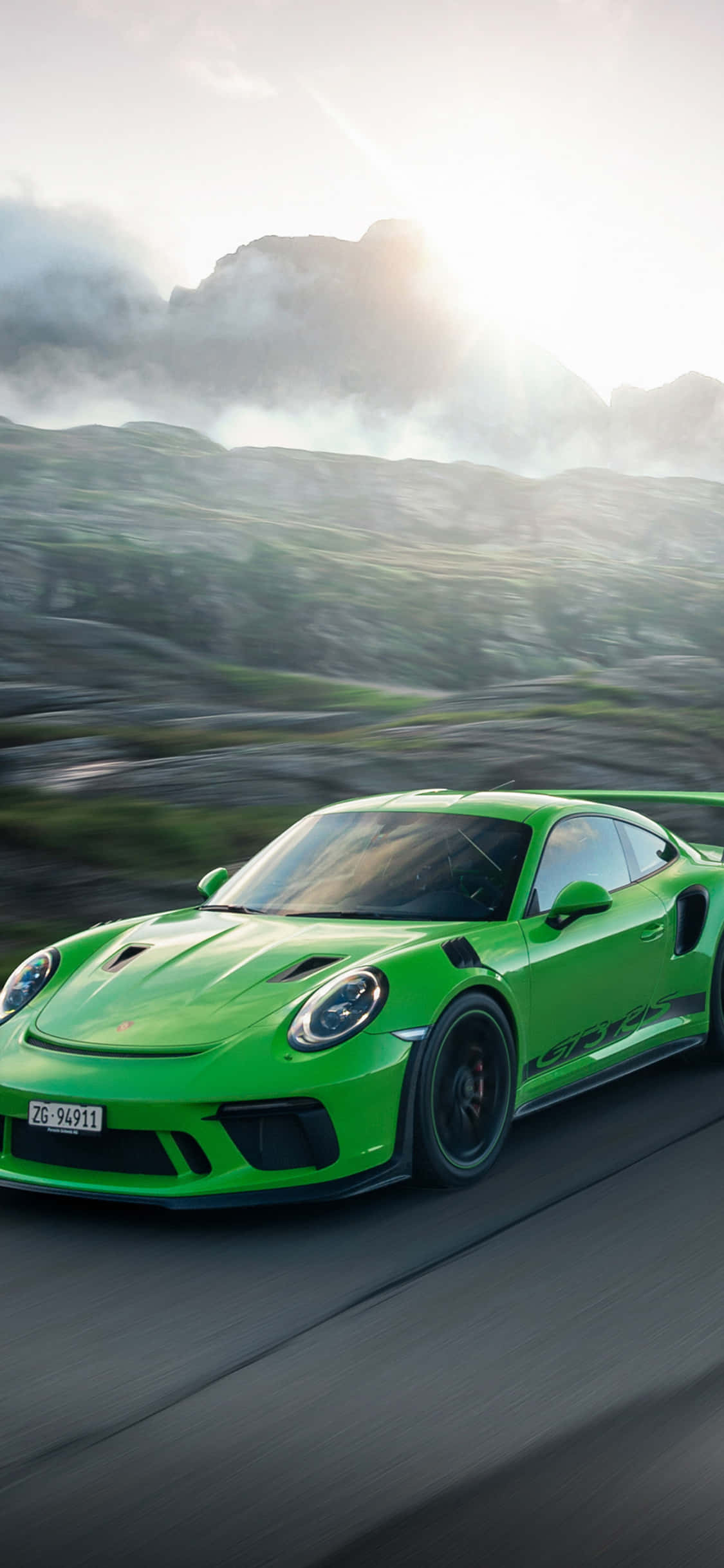 Porsche iPhone 桌布 壁紙 — 一輛充滿活力的綠色保時捷 911 GT2 RS 在蜿蜒的山路上疾馳，背景是動態模糊和陽光普照的山峰。