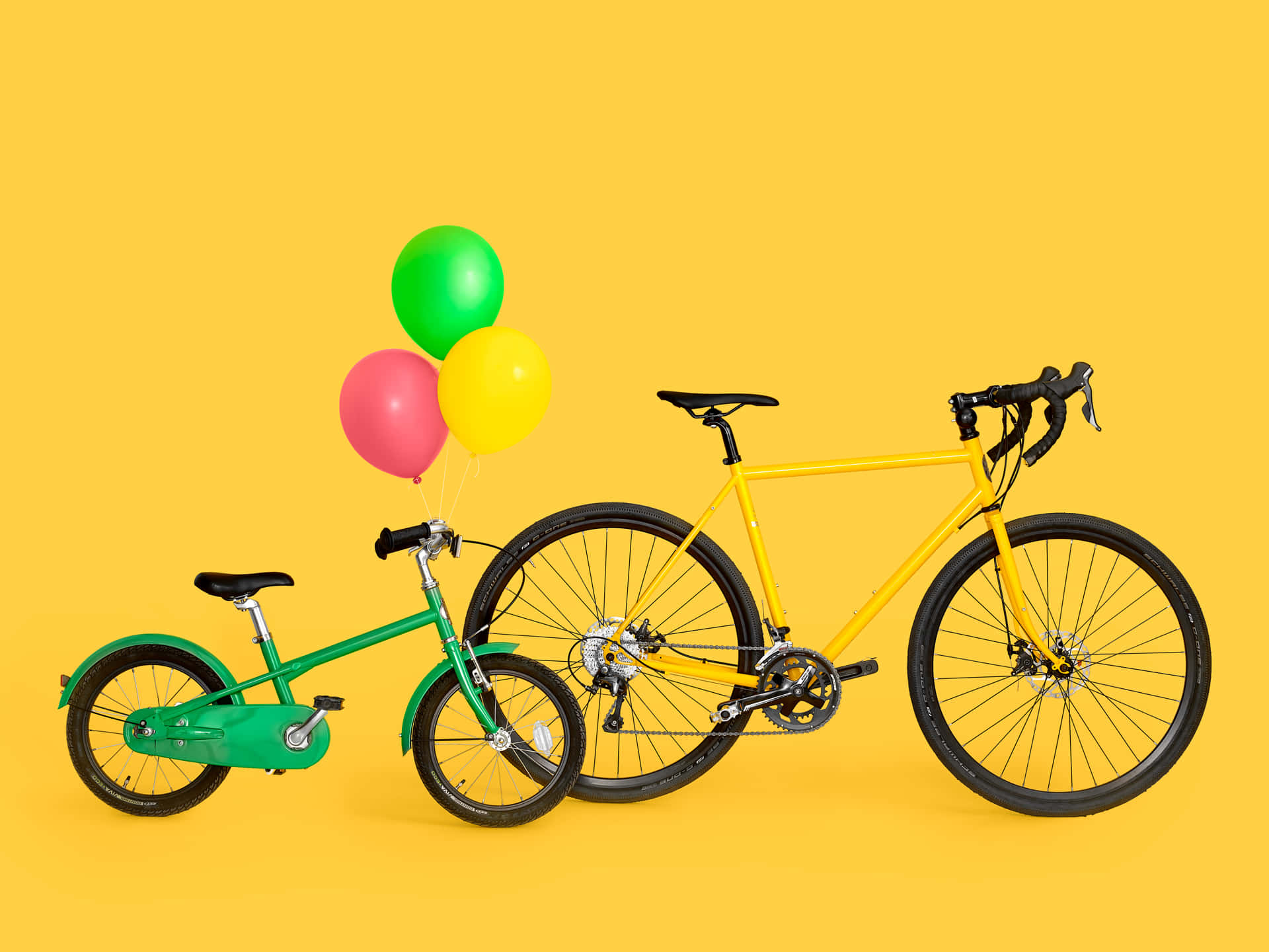 Balloons 桌布 壁紙 — 一輛充滿活力的綠色兒童腳踏車和黃色成人腳踏車並排立在純黃色背景前，上方有三個彩色氣球飄浮。