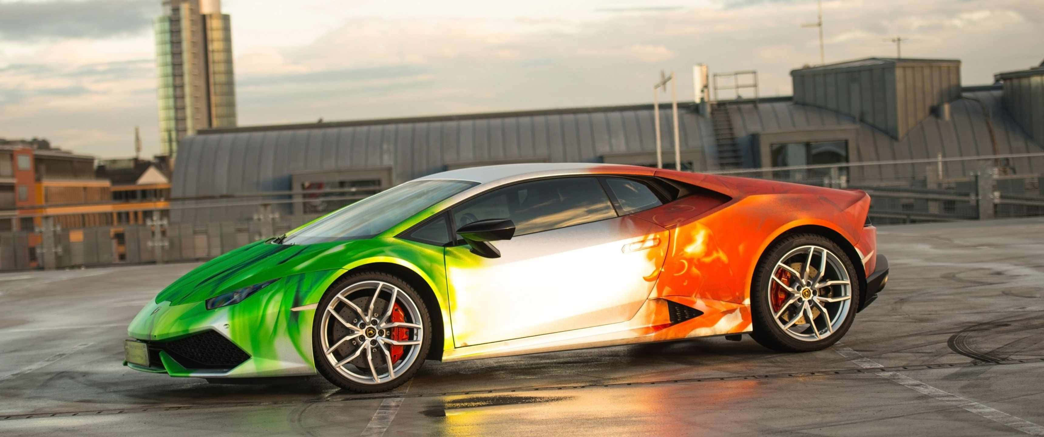 3440x1440p Lamborghini 桌布 壁紙 — 一輛充滿活力的綠色和橙色藍寶堅尼 Huracán 停泊在屋頂上，背景是日落時分的城市建築。