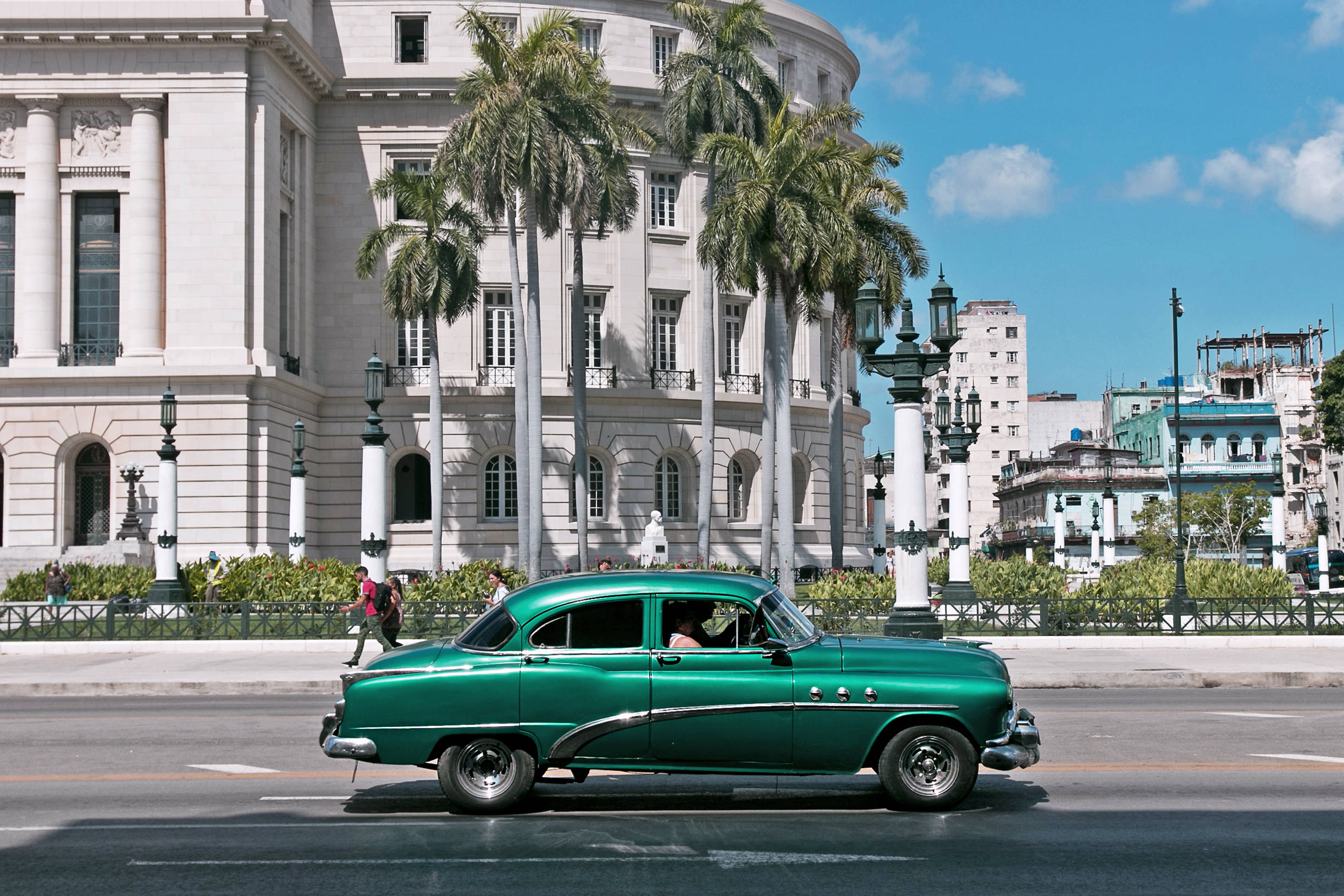 Cuba 桌布 壁紙 — 一輛充滿活力的綠色復古車駛過一座宏偉的新古典主義建築，天空湛藍，點綴著蓬鬆的白雲。