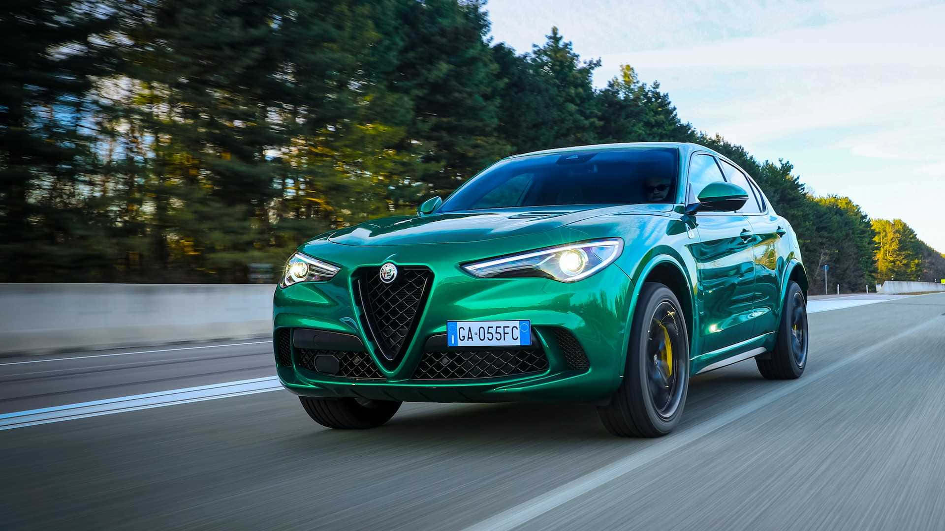 Alfa Romeo Stelvio 桌布 壁紙 — 一輛充滿活力的綠色愛快羅密歐Stelvio SUV在樹木成蔭的道路上奔馳，動態模糊傳達出動感。