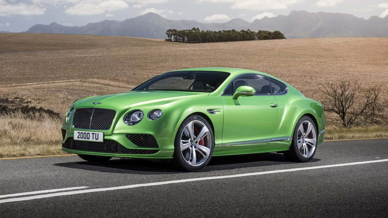 賓利 桌布 壁紙 — 一輛充滿活力的綠色賓利 Continental GT 雙門跑車在鄉村道路上巡航，周圍是乾燥的田野和遠處的山脈，天空部分多雲。