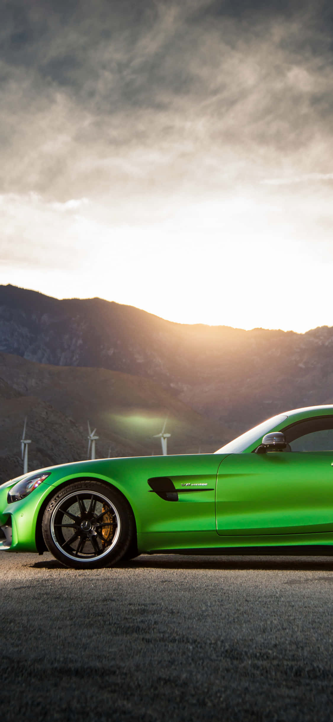 iPhone X Amg Gt-r 桌布 壁紙 — 一輛充滿活力的綠色跑車停在瀝青路上，背景有風力發電機和山脈，天空呈現金黃色的日落。