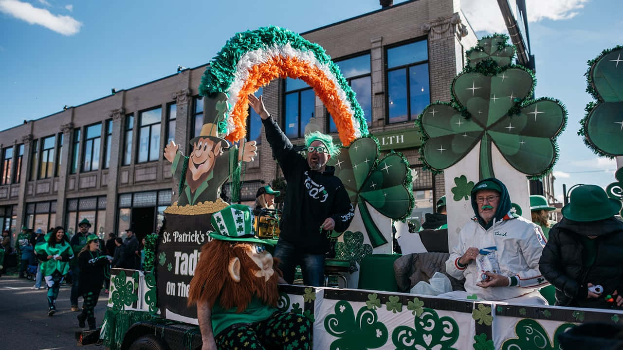 St Patrick's Day 桌布 壁紙 — 一輛充滿活力的聖派翠克節遊行花車，裝飾著綠色三葉草和一道彩虹，拱形在穿著節日服裝的歡呼狂歡者上方。
