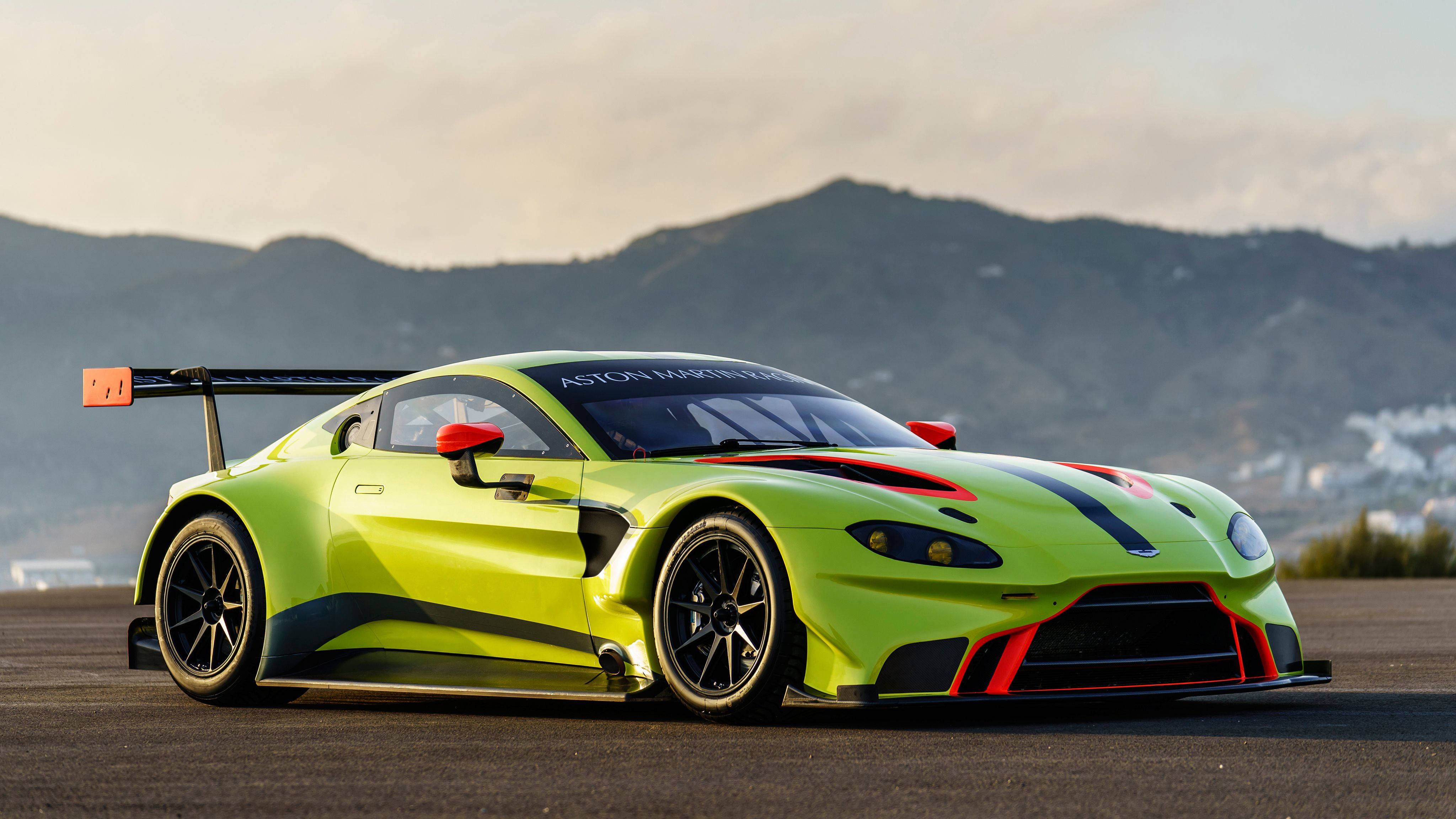 奧斯頓馬丁 桌布 壁紙 — 一輛充滿活力的萊姆綠色 Aston Martin 賽車，帶有紅色點綴和大型後翼，停在瀝青賽道上，背景是山脈。