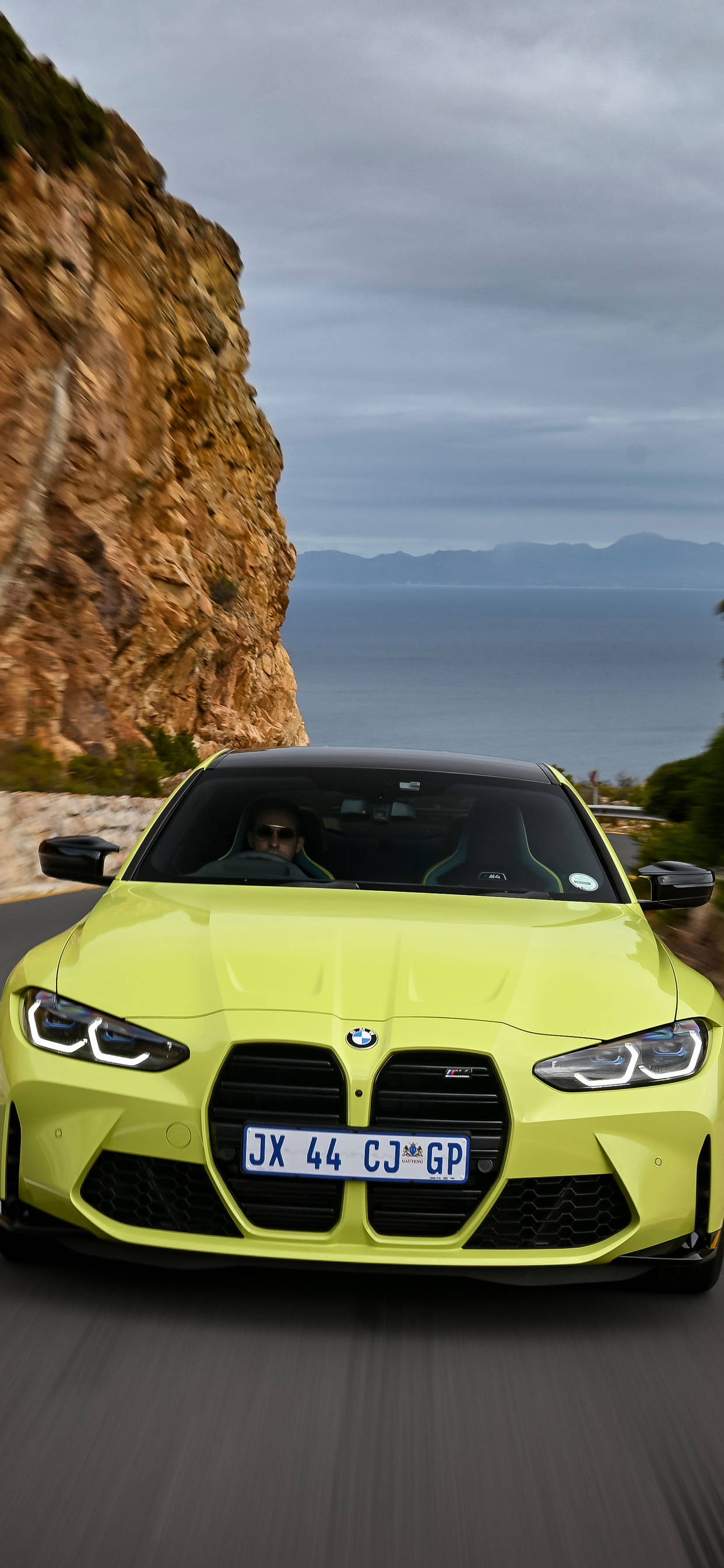 m 桌布 壁紙 — 一輛充滿活力的萊姆綠色BMW M4沿著海岸懸崖道路疾馳，海洋和遠處的山脈在陰天之下。