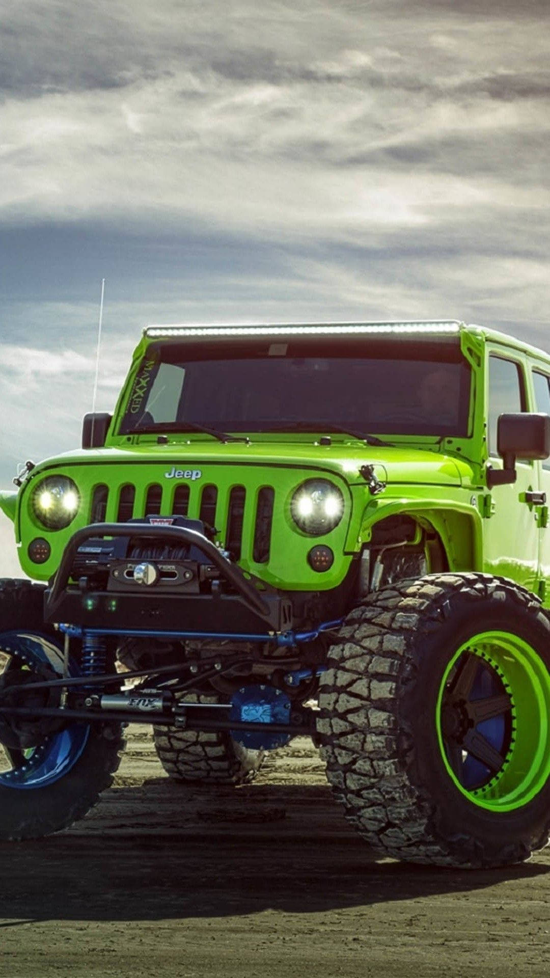 吉普 桌布 壁紙 — 一輛充滿活力的萊姆綠色Jeep Wrangler，配備超大輪胎和藍色懸吊，站在多塵的地形上，天空多雲。