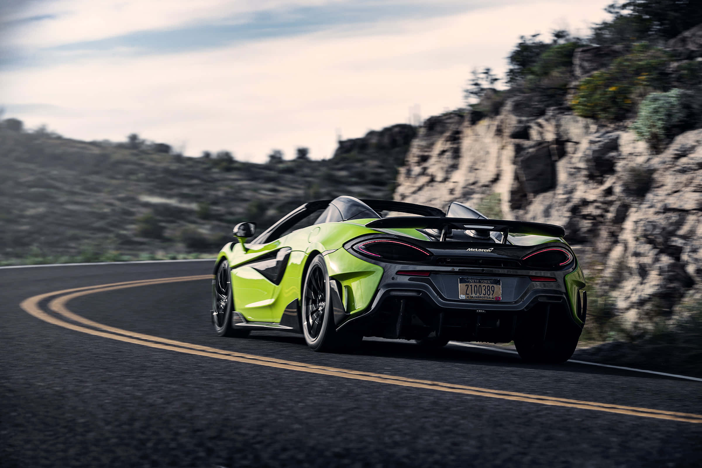 Mclaren 600lt Spider 桌布 壁紙 — 一輛充滿活力的萊姆綠色McLaren跑車在蜿蜒的山路上奔馳，兩旁是岩石峭壁和稀疏的植被，天空多雲。