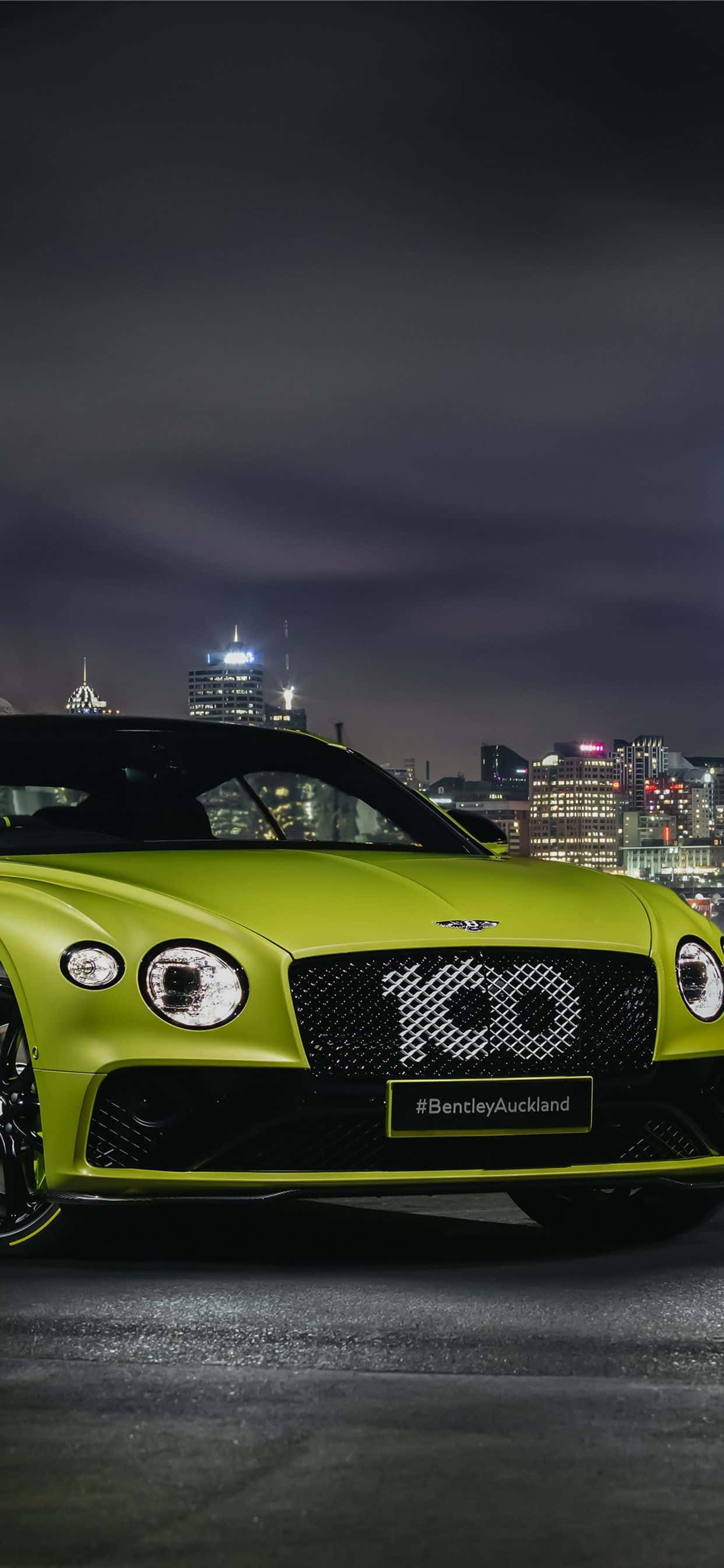 iPhone Xs Max Bentley 桌布 壁紙 — 一輛充滿活力的萊姆綠色賓利跑車在夜燈下閃耀，映襯著發光的城市天際線。