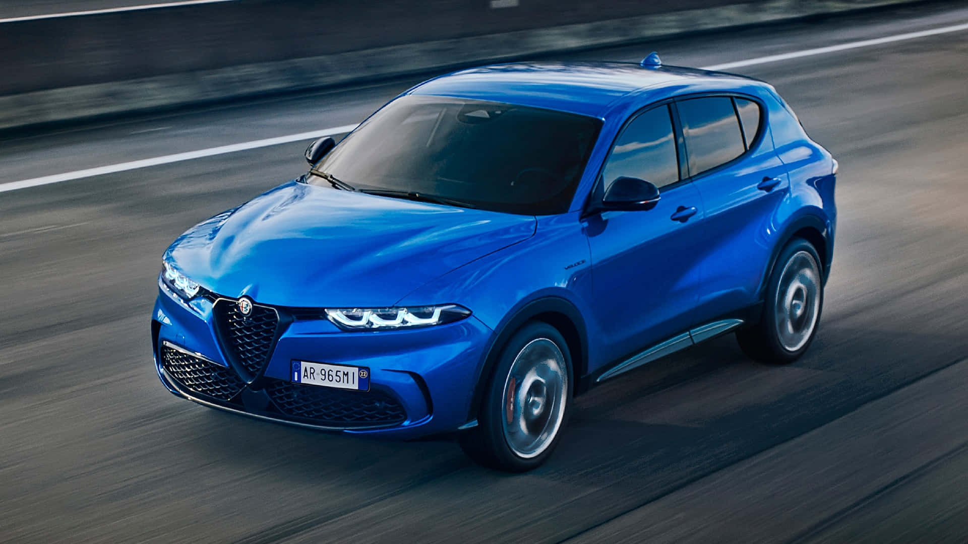 愛快羅密歐 桌布 壁紙 — 一輛充滿活力的藍色 Alfa Romeo SUV 在模糊的高速公路上疾馳，動態模糊強調了其動態運動。