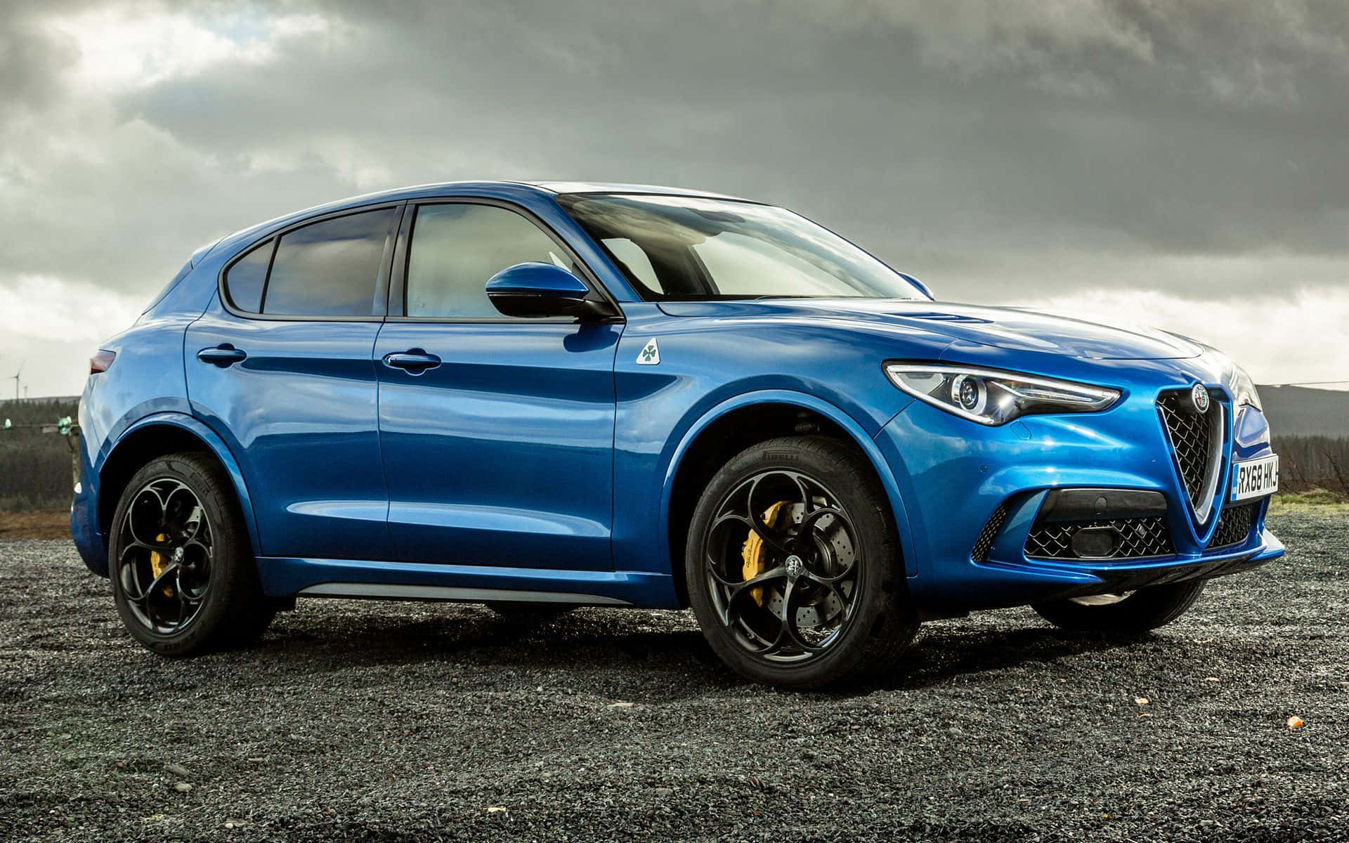 Alfa Romeo Stelvio 桌布 壁紙 — 一輛充滿活力的藍色 Alfa Romeo SUV，配備黑色車輪和黃色制動卡鉗，停放在碎石地上，天空多雲。
