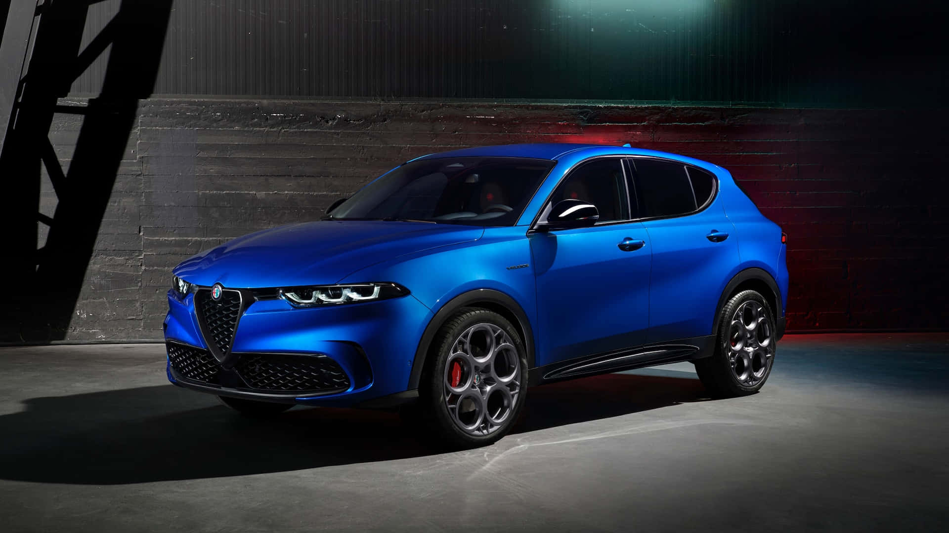 愛快羅密歐 桌布 壁紙 — 一輛充滿活力的藍色 Alfa Romeo SUV 在戲劇性的攝影棚燈光下，在深色紋理牆壁前閃閃發光。