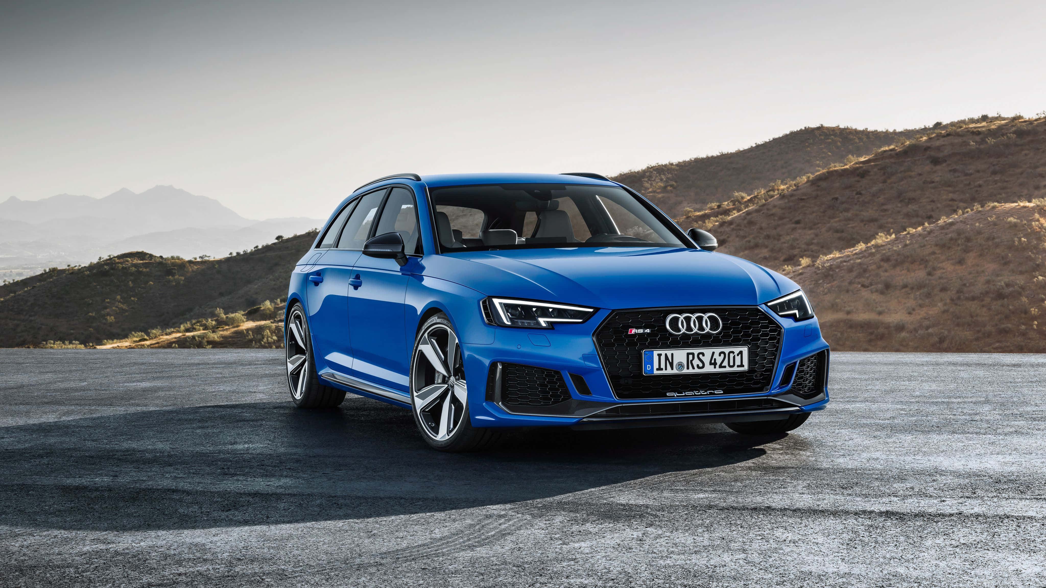 Audi Rs4 桌布 壁紙 — 一輛充滿活力的藍色 Audi RS4 Avant 旅行車停放在鋪砌道路上，背景是乾燥的山丘和遠處的山脈，天空朦朧。