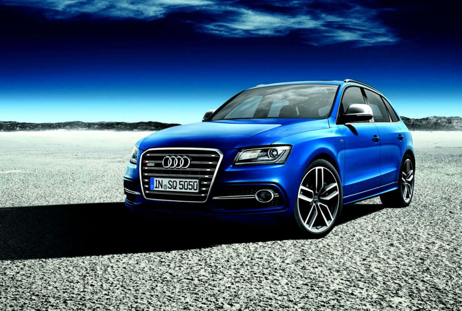 Audi Sq5 桌布 壁紙 — 一輛充滿活力的藍色 Audi SQ5 SUV 大膽地矗立在礫石平原上，背景是帶有縷縷雲彩的戲劇性深藍色天空。