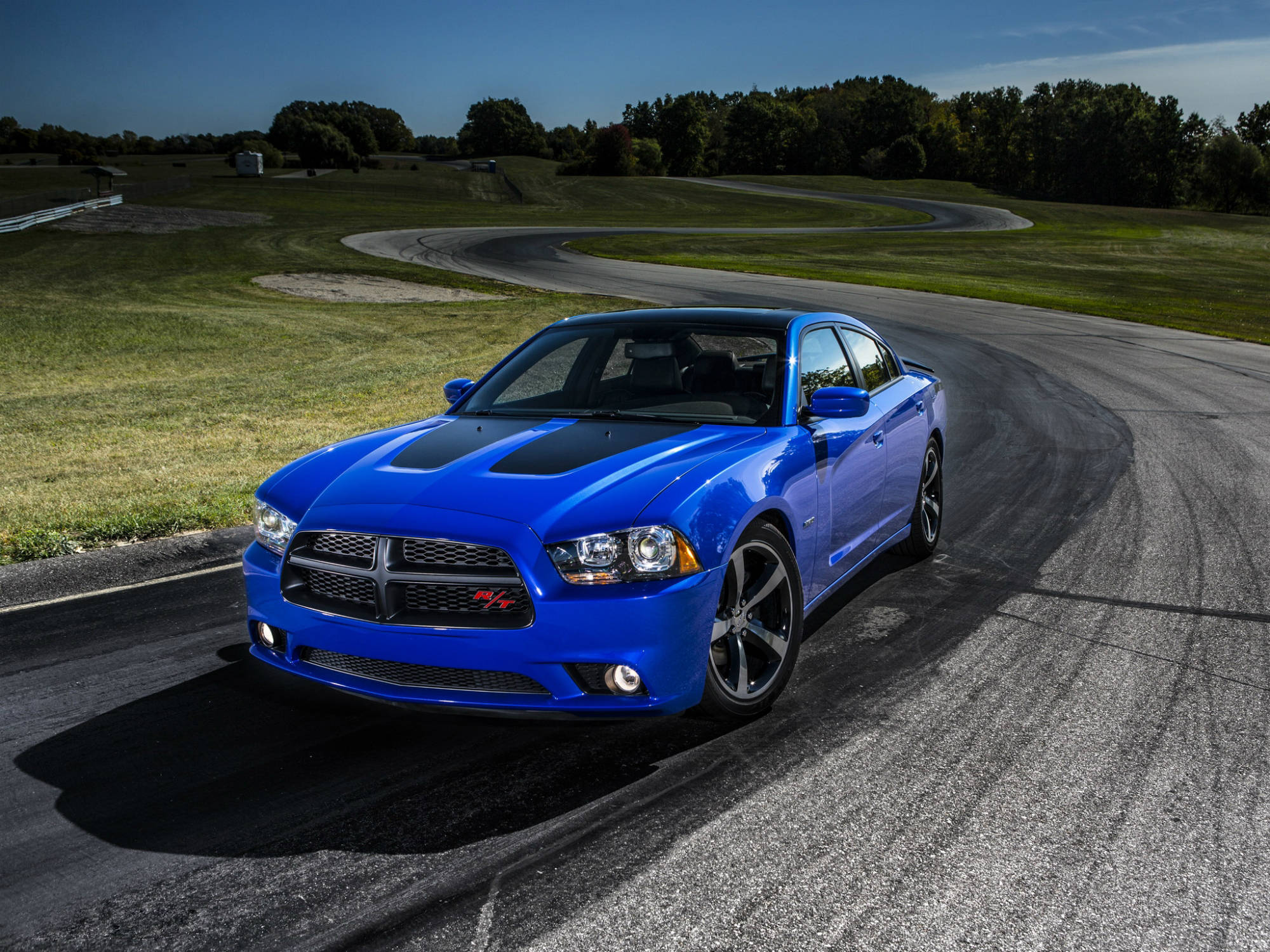 道奇隊 桌布 壁紙 — 一輛充滿活力的藍色 Dodge Charger R/T 在彎曲的瀝青賽道上疾馳，周圍環繞著茂密的綠草和樹木，天空清澈湛藍。
