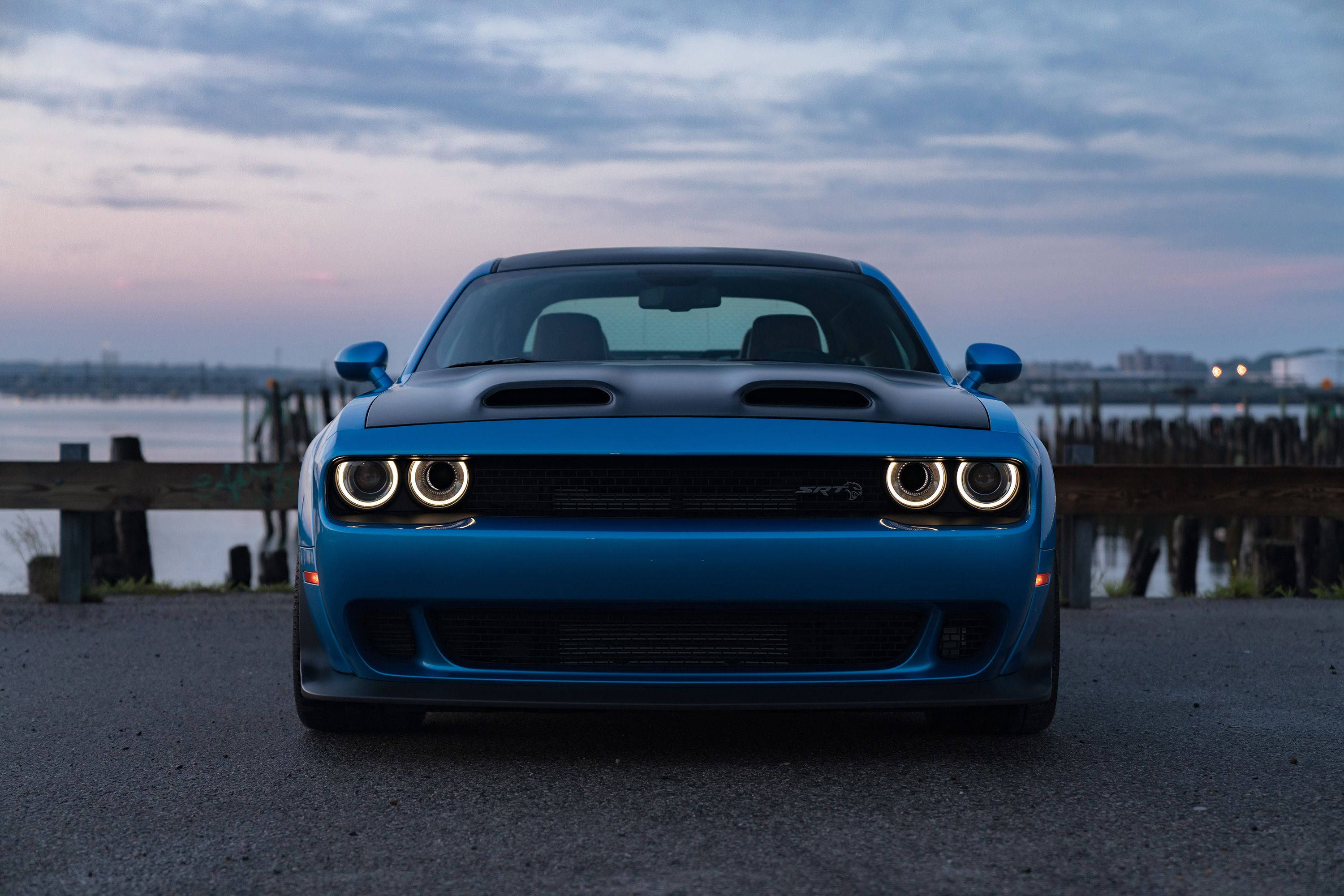 Cool Car 桌布 壁紙 — 一輛充滿活力的藍色 Dodge Challenger SRT Hellcat 正面朝前，圓形頭燈發光，背景是暮色中的海濱。