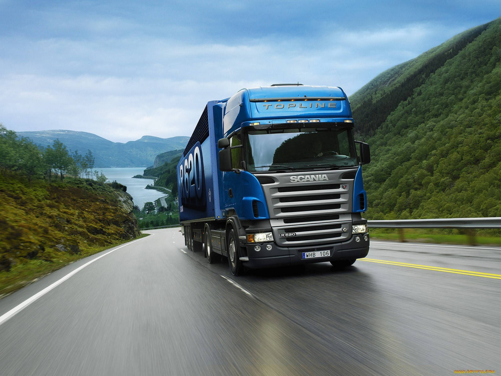 Trucks 桌布 壁紙 — 一輛充滿活力的藍色 Scania 卡車在陰天之下，沿著蜿蜒的山路在寧靜的湖邊奔馳。