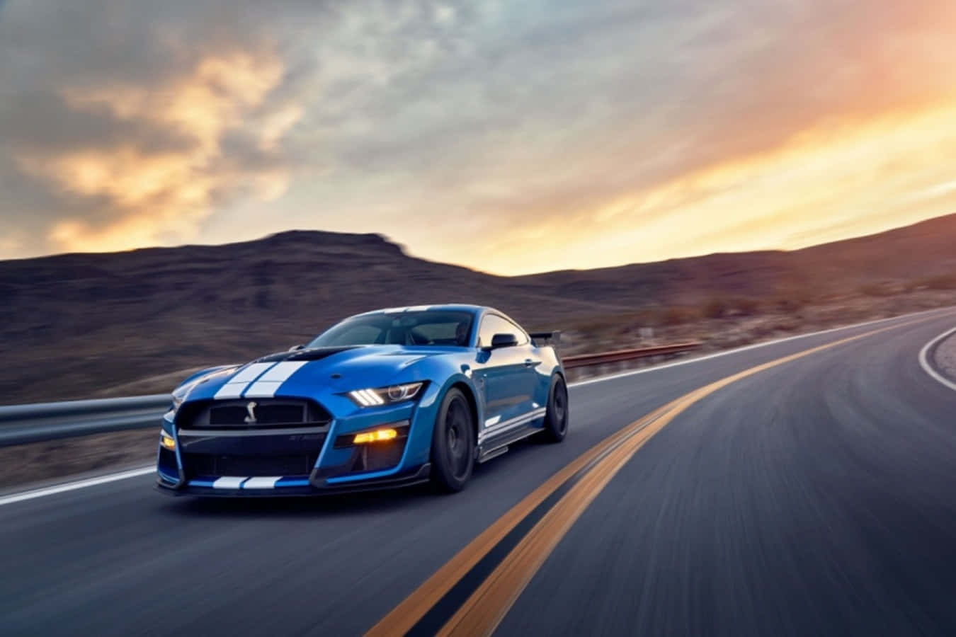 野馬GIF 桌布 壁紙 — 一輛充滿活力的藍色 Shelby Mustang GT500 在日落時分沿著蜿蜒的沙漠高速公路疾馳，頭頂上有戲劇性的雲彩。