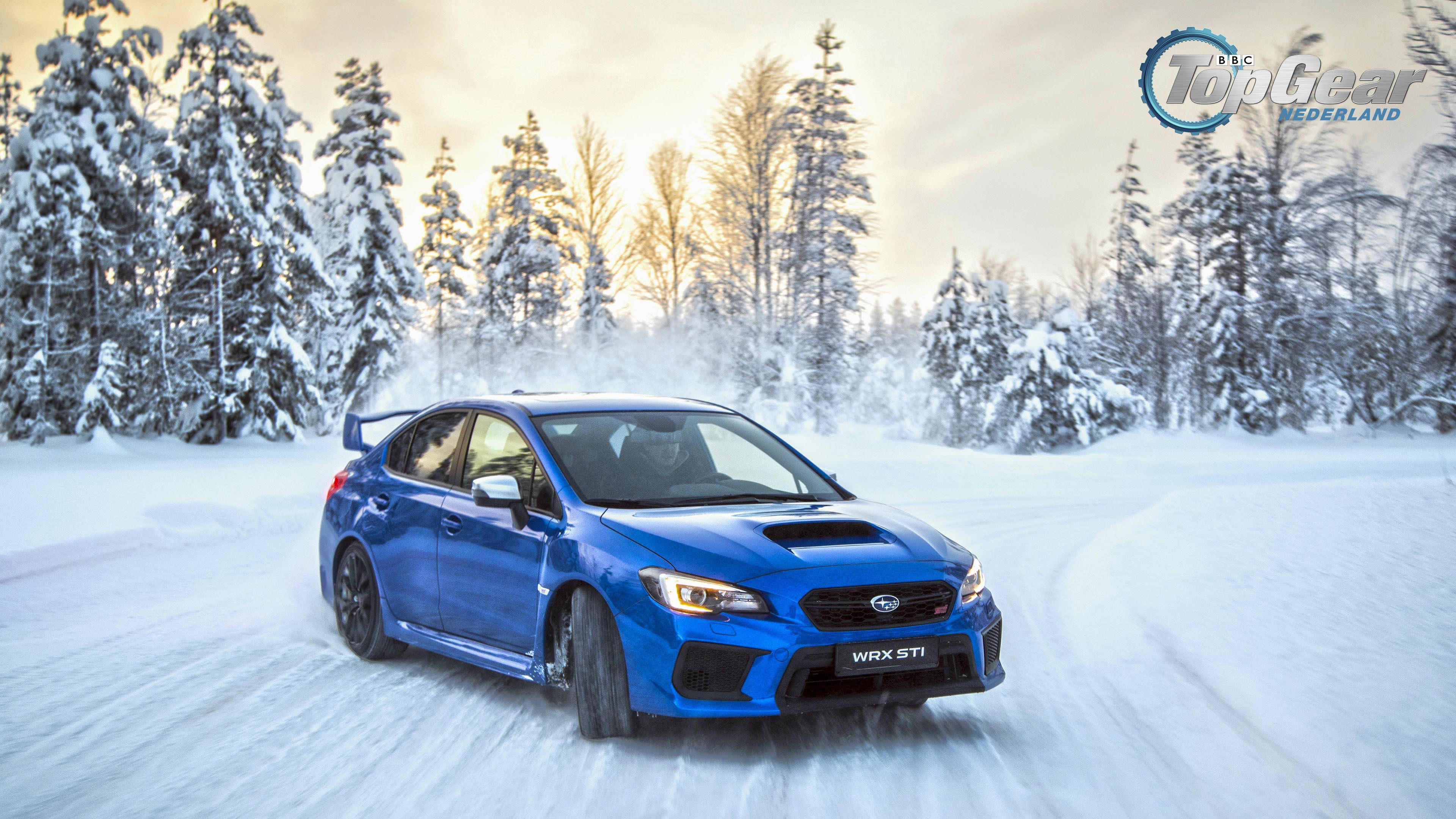 昴 桌布 壁紙 — 一輛充滿活力的藍色 Subaru WRX STI 在雪地森林中疾馳，輪胎濺起雪花，天空呈現柔和的金色。