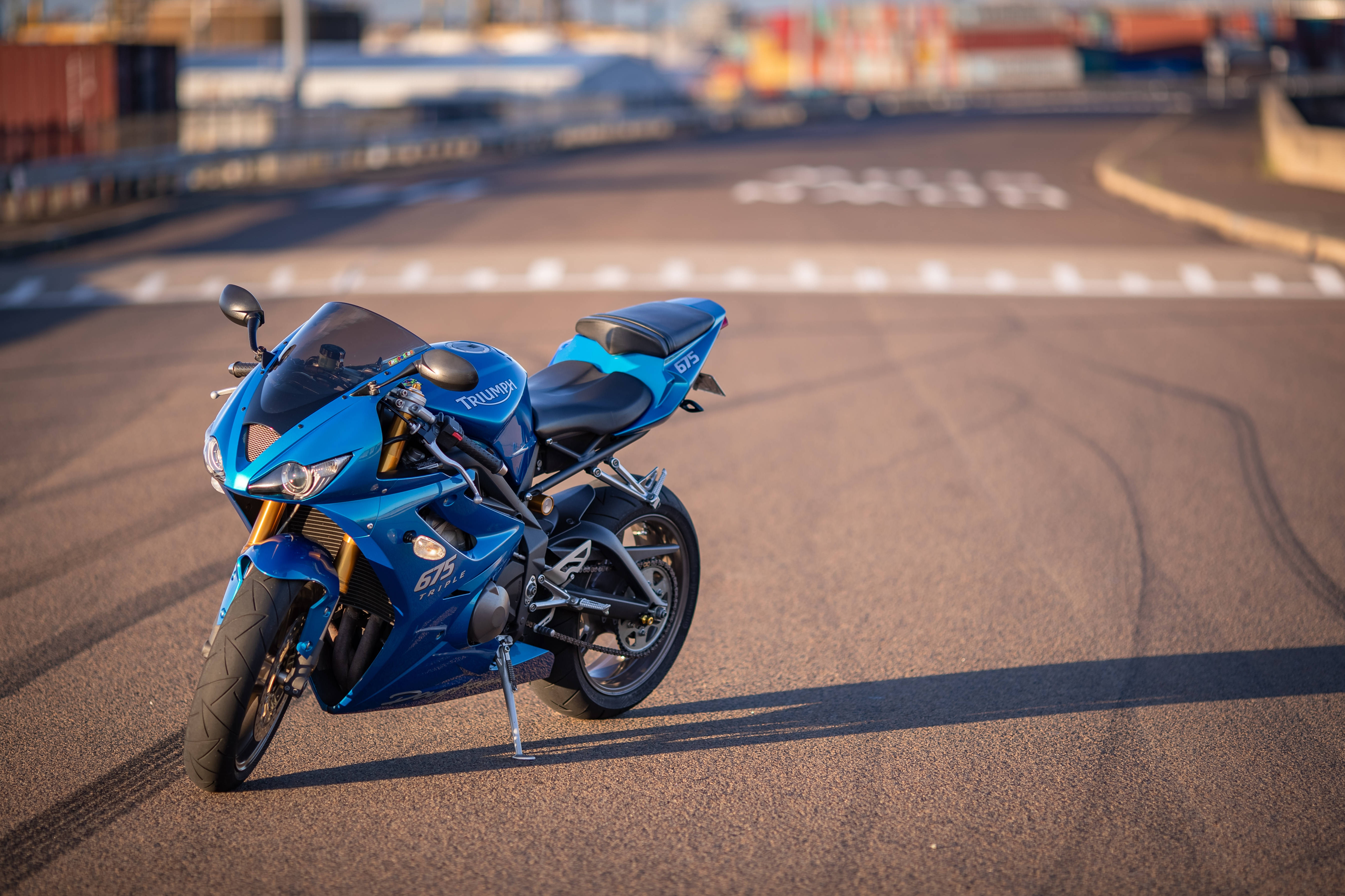  壁紙 — 一輛充滿活力的藍色 Triumph 摩托車停放在陽光普照的賽道上，背景有輪胎痕跡和模糊的貨運集裝箱。