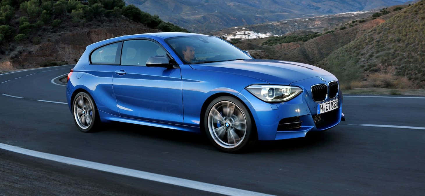 Bmw 1 Series 桌布 壁紙 — 一輛充滿活力的藍色BMW 1系列掀背車沿著蜿蜒的山路疾馳，背景是崎嶇的山丘和遠處的村莊。