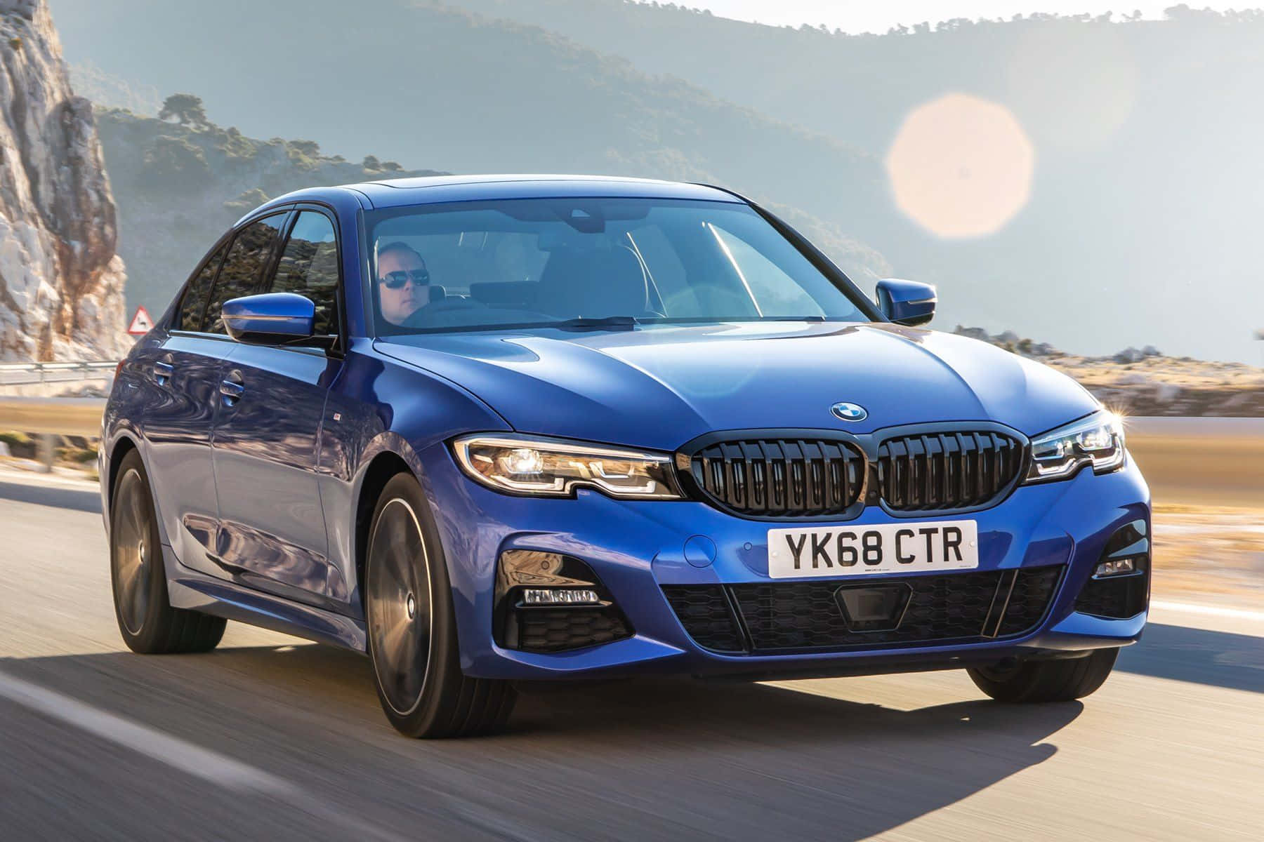 Bmw 3 Series 桌布 壁紙 — 一輛充滿活力的藍色BMW 3系列轎車沿著海岸公路疾馳，背景有太陽耀斑和岩石峭壁。