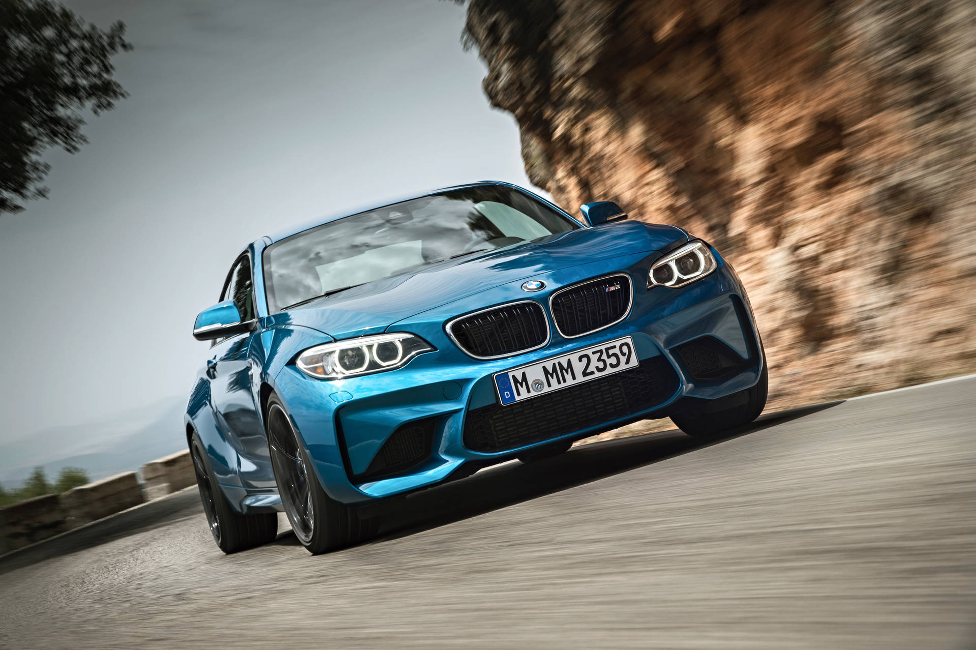 桌布 壁紙 — 一輛充滿活力的藍色BMW M2 Coupe沿著蜿蜒的山路疾馳，帶有模糊的動態效果，旁邊是岩石峭壁。