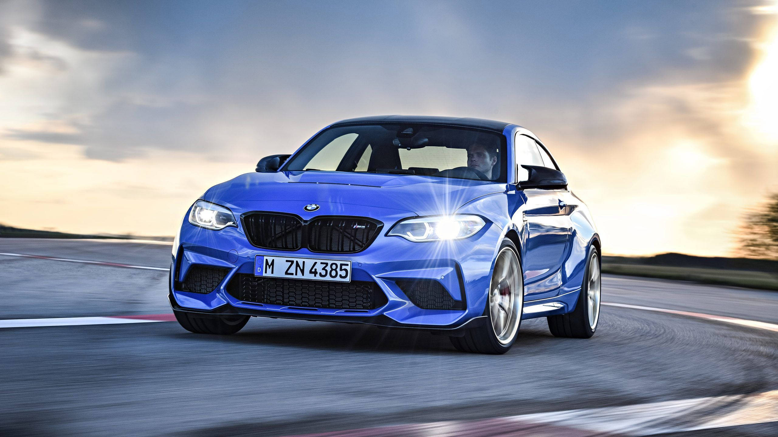 Bmw Cars 桌布 壁紙 — 一輛充滿活力的藍色BMW M2 Coupe在日落時分沿著賽道疾馳，車頭燈閃爍，帶有動態模糊效果。
