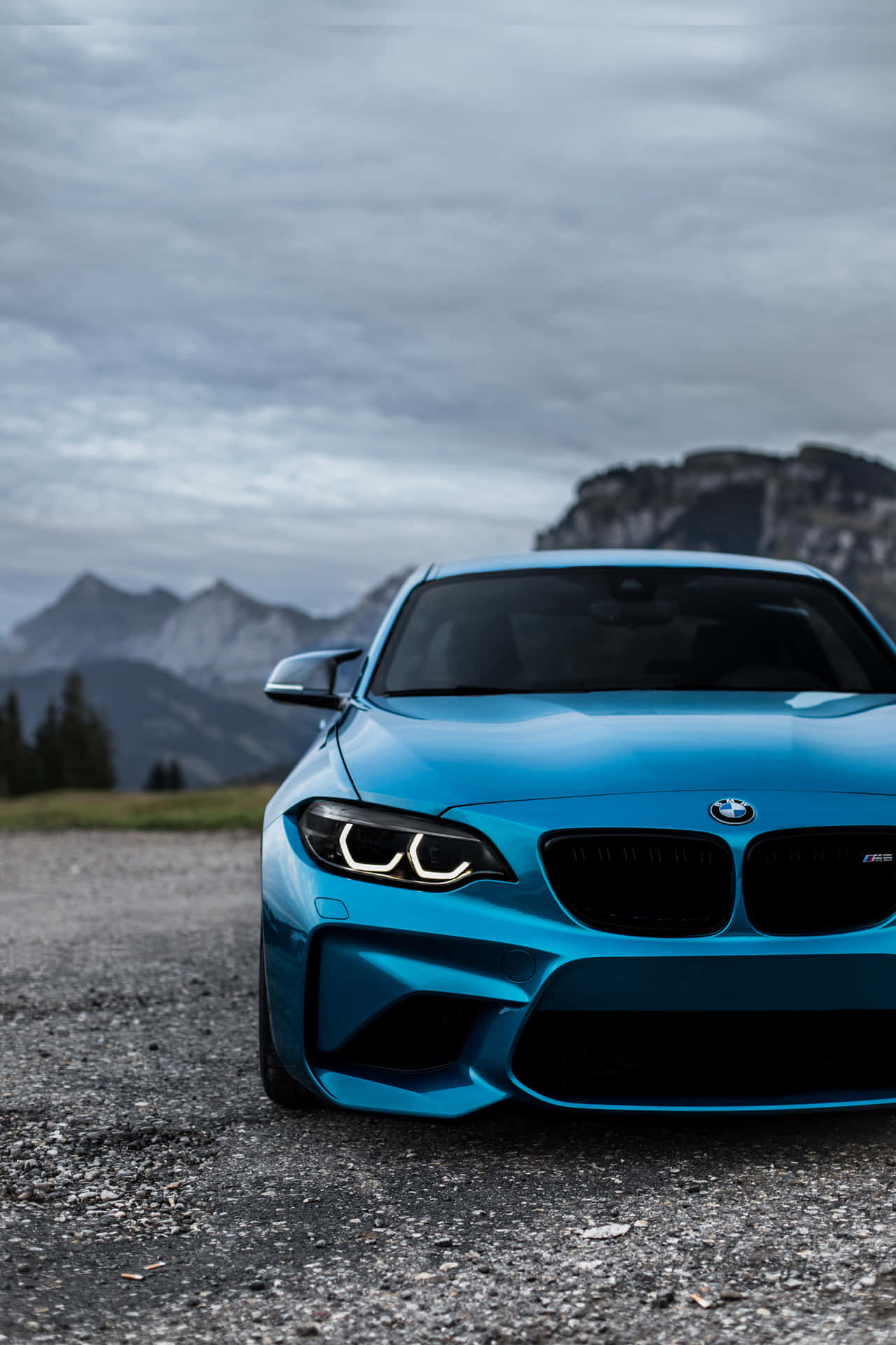 BMW 動圖 桌布 壁紙 — 一輛充滿活力的藍色BMW M2跑車停在礫石上，背景是陰雲密布的天空下戲劇性的山頂。