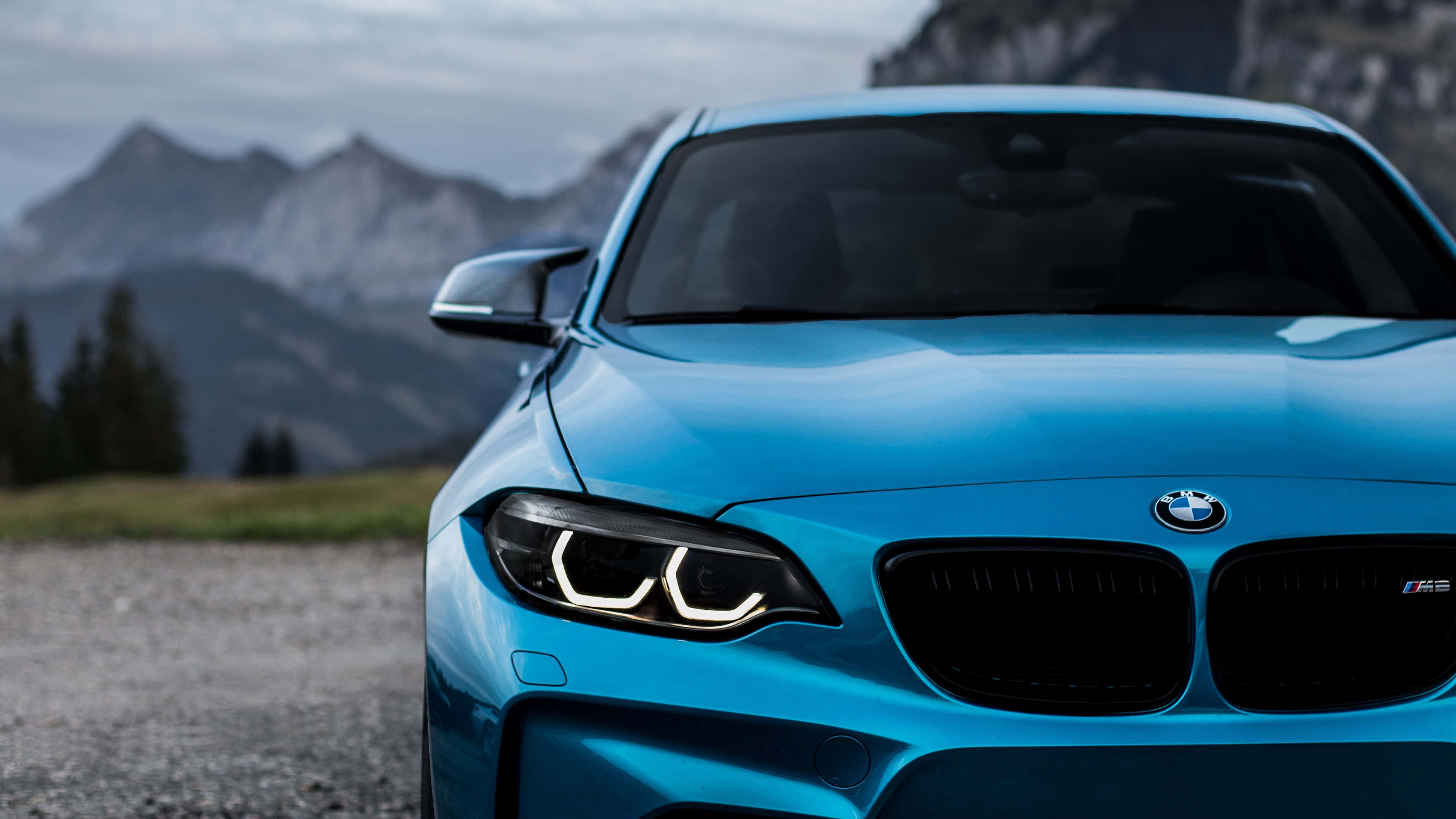 BMW 動圖 桌布 壁紙 — 一輛充滿活力的藍色BMW M2跑車停在碎石路上，背景是霧濛濛的山脈。