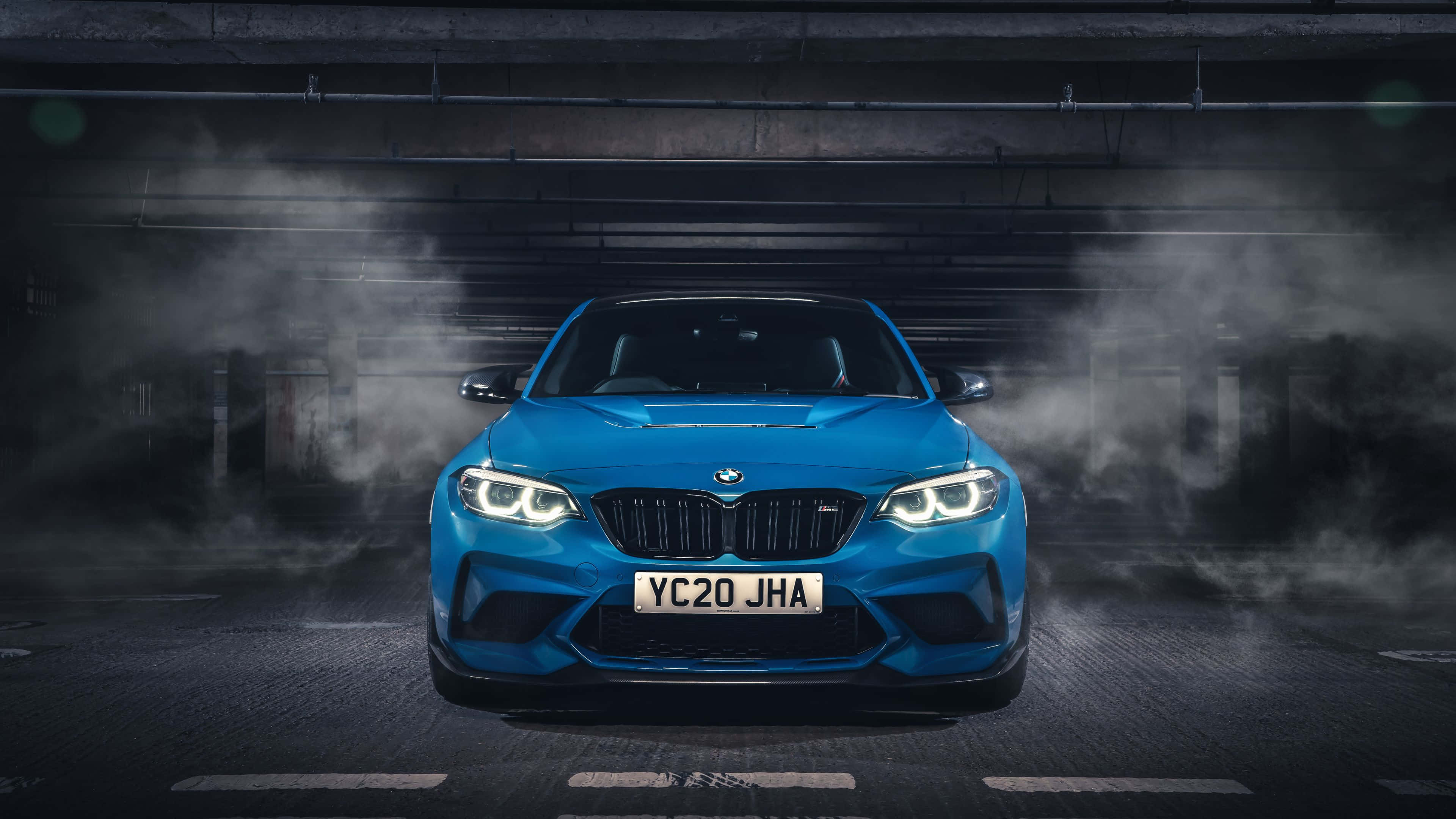 4K BMW 壁紙 桌布 壁紙 — 一輛充滿活力的藍色BMW M2跑車，閃爍的車頭燈主導了煙霧瀰漫的地下停車場。