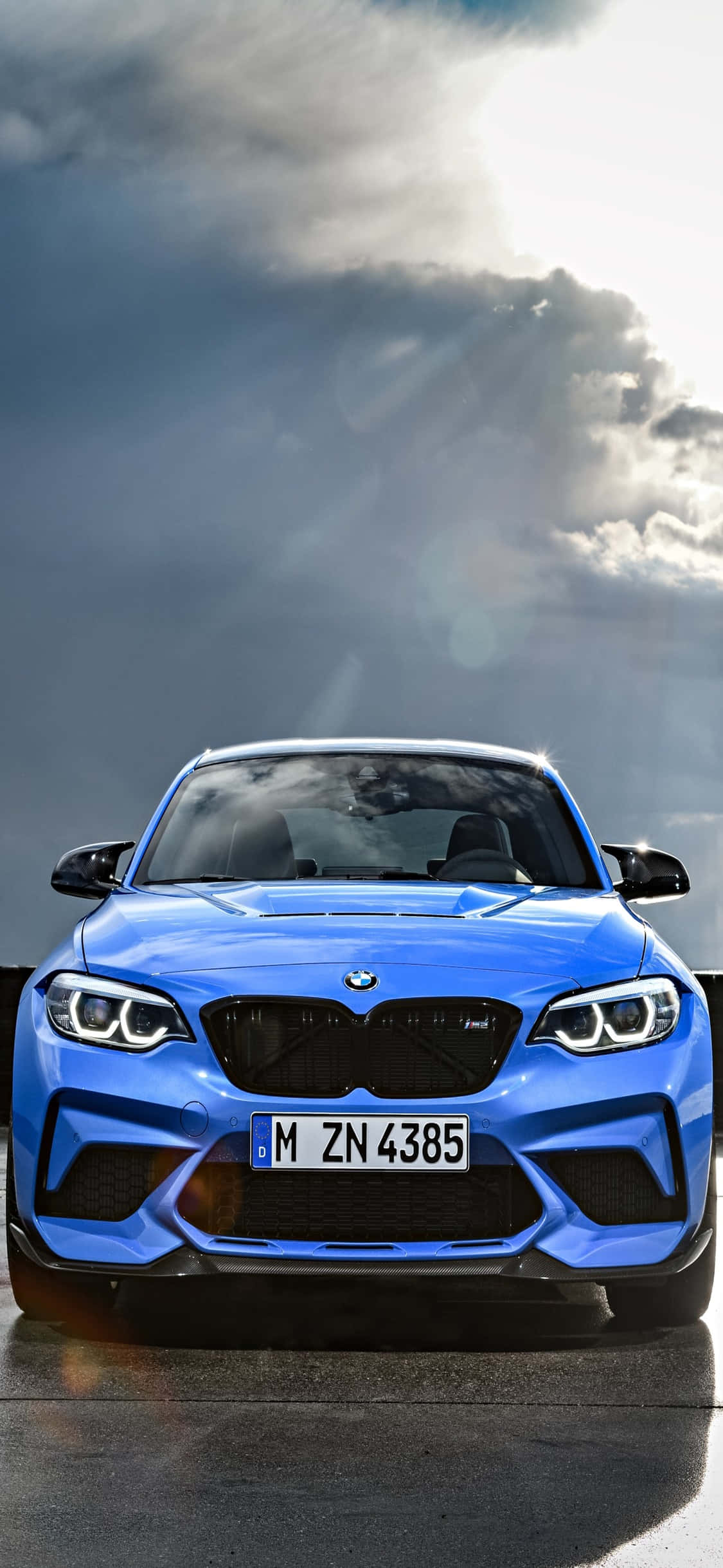 iPhone X Bmw 桌布 壁紙 — 一輛充滿活力的藍色BMW M2跑車在戲劇性的多雲天空下，陽光穿透雲層，正面朝前。