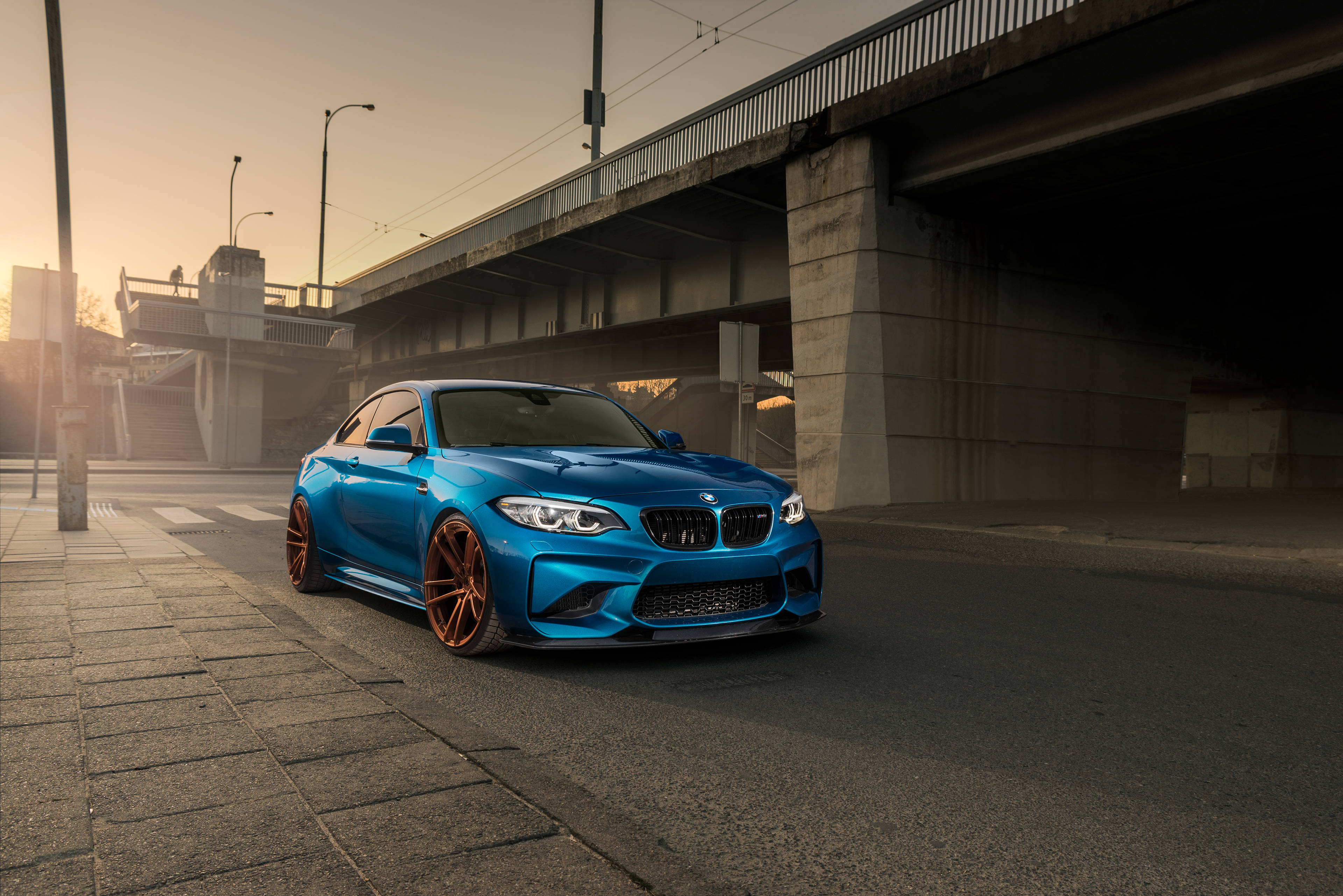 4K BMW 壁紙 桌布 壁紙 — 一輛充滿活力的藍色BMW M2雙門跑車，配備銅色輪圈，停泊在混凝土高架橋下，正值黃金時刻。