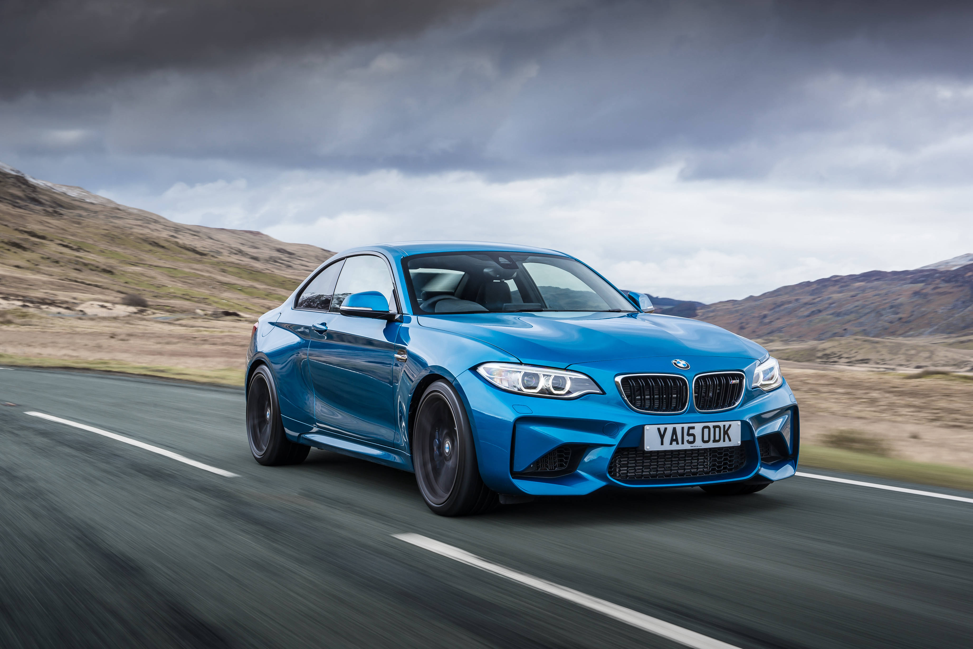 4K BMW 壁紙 桌布 壁紙 — 一輛充滿活力的藍色BMW M2雙門跑車，在戲劇性的多雲天空下，沿著蜿蜒的道路穿過崎嶇的山丘疾馳。