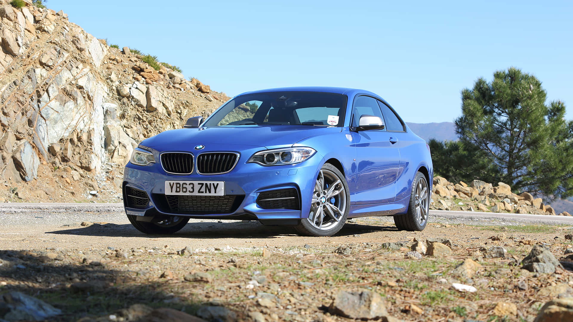 Bmw 2 Series 桌布 壁紙 — 一輛充滿活力的藍色BMW M2雙門跑車停泊在岩石路邊，天空清澈湛藍，樹木稀疏，遠處有山丘。