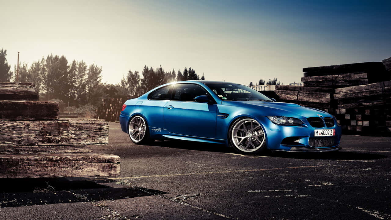 720p Bmw 桌布 壁紙 — 一輛充滿活力的藍色BMW M3雙門跑車在黃金時段的光線下閃閃發光，停放在堆疊的木樑旁邊。