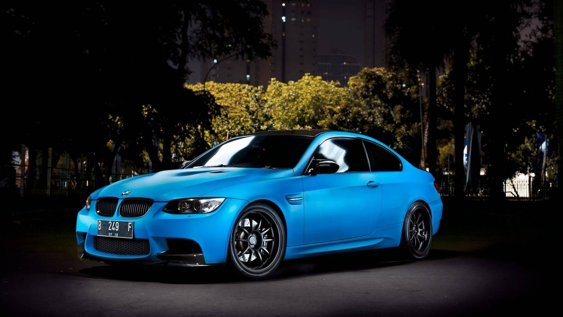 藍色 桌布 壁紙 — 一輛充滿活力的藍色BMW M3雙門轎跑車在夜間城市燈光下閃閃發光，背景有樹木和建築物。