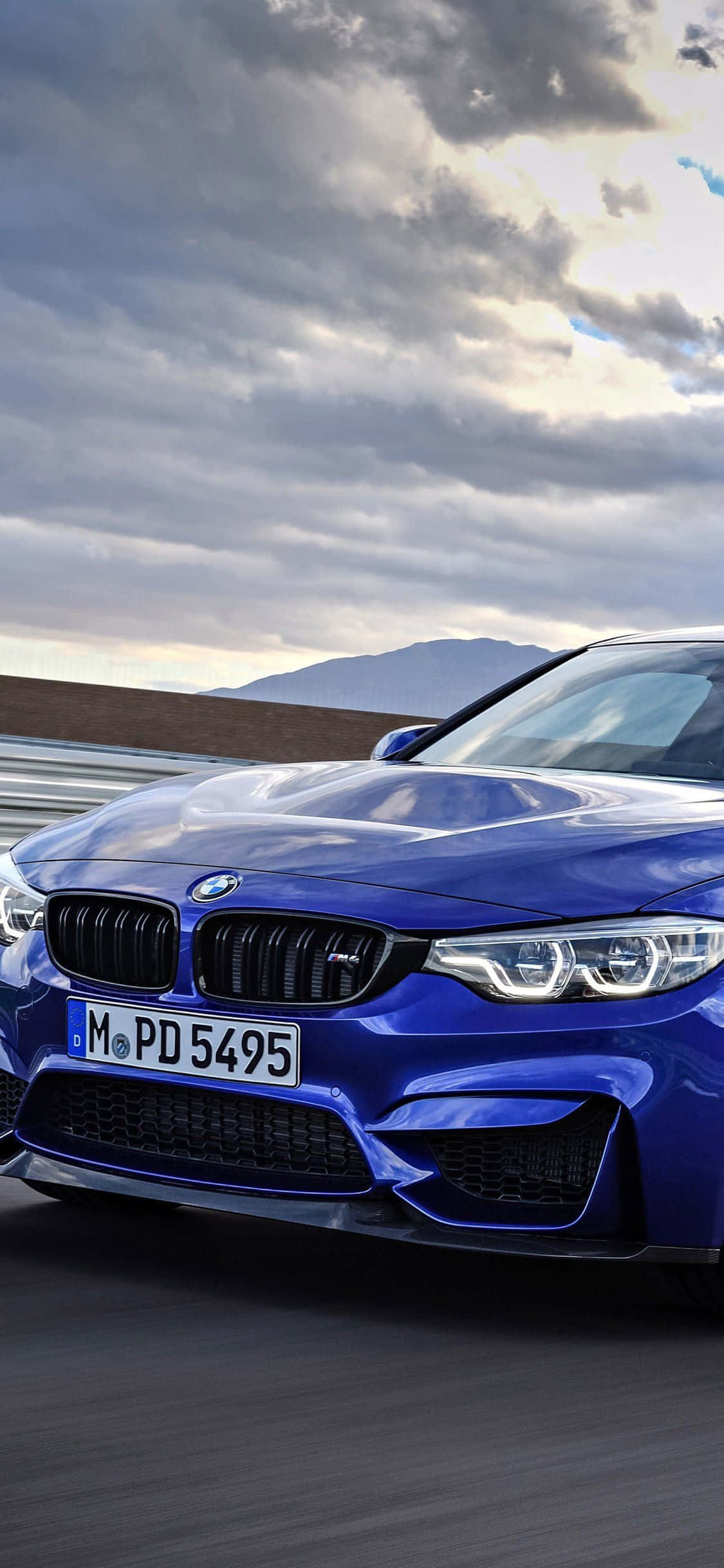 Bmw iPhone 桌布 壁紙 — 一輛充滿活力的藍色BMW M4跑車在戲劇性的多雲天空下沿著山路飛馳。