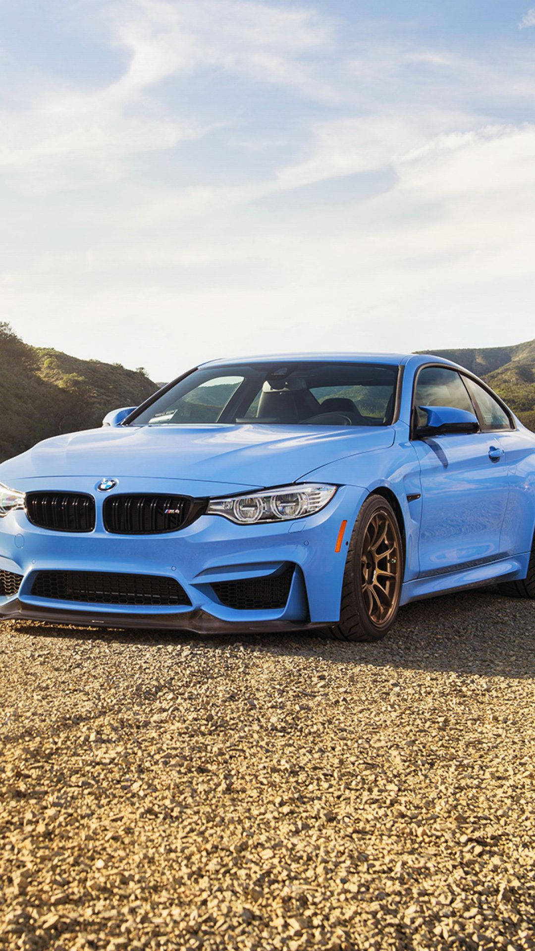 BMW 動圖 桌布 壁紙 — 一輛充滿活力的藍色BMW M4雙門跑車停在碎石上，背景是起伏的丘陵，天空部分多雲。