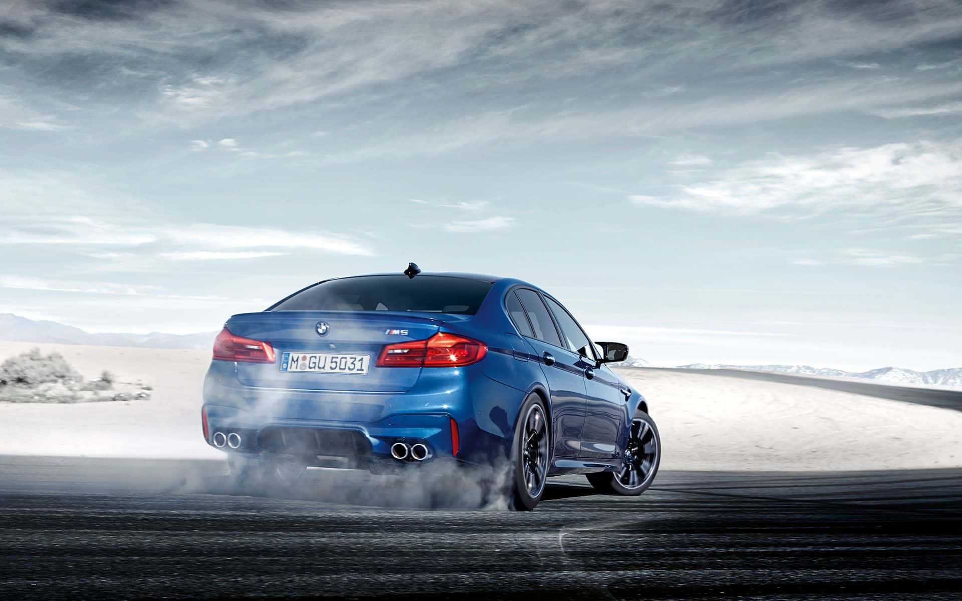 BMW 動圖 桌布 壁紙 — 一輛充滿活力的藍色BMW M5在沙漠道路上飄移，在陰天天空下揚起煙霧。