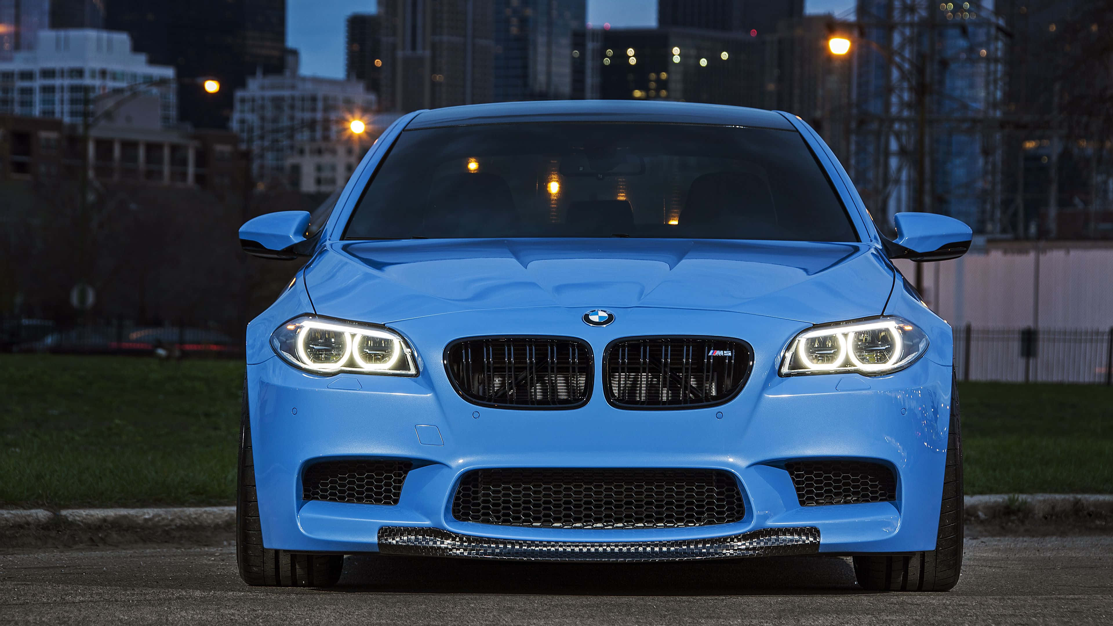 桌布 壁紙 — 一輛充滿活力的藍色BMW M5跑車在暮色城市燈光下閃耀，車頭燈亮起。
