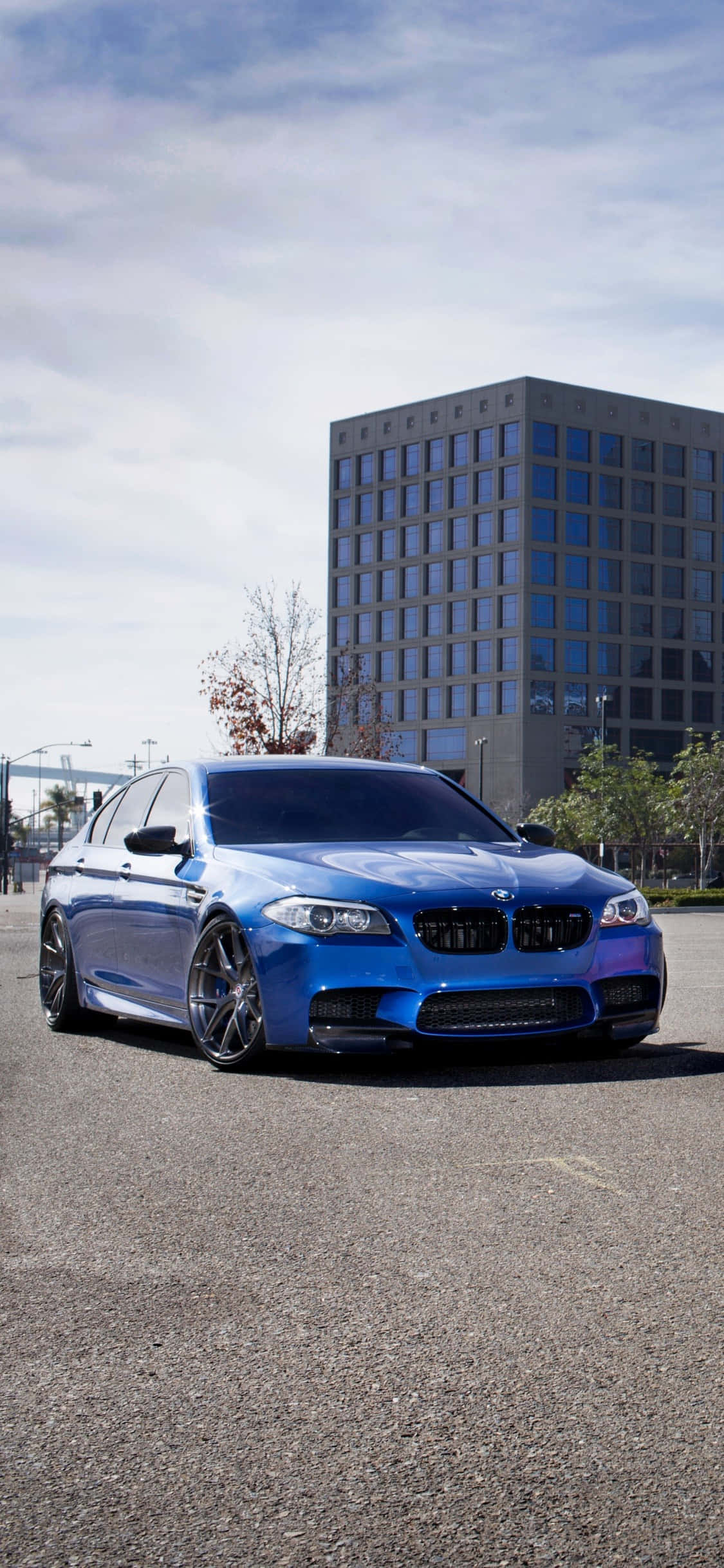 iPhone X Bmw 桌布 壁紙 — 一輛充滿活力的藍色BMW M5跑車停在瀝青路上，旁邊有現代玻璃建築和稀疏的樹木，天空部分多雲。