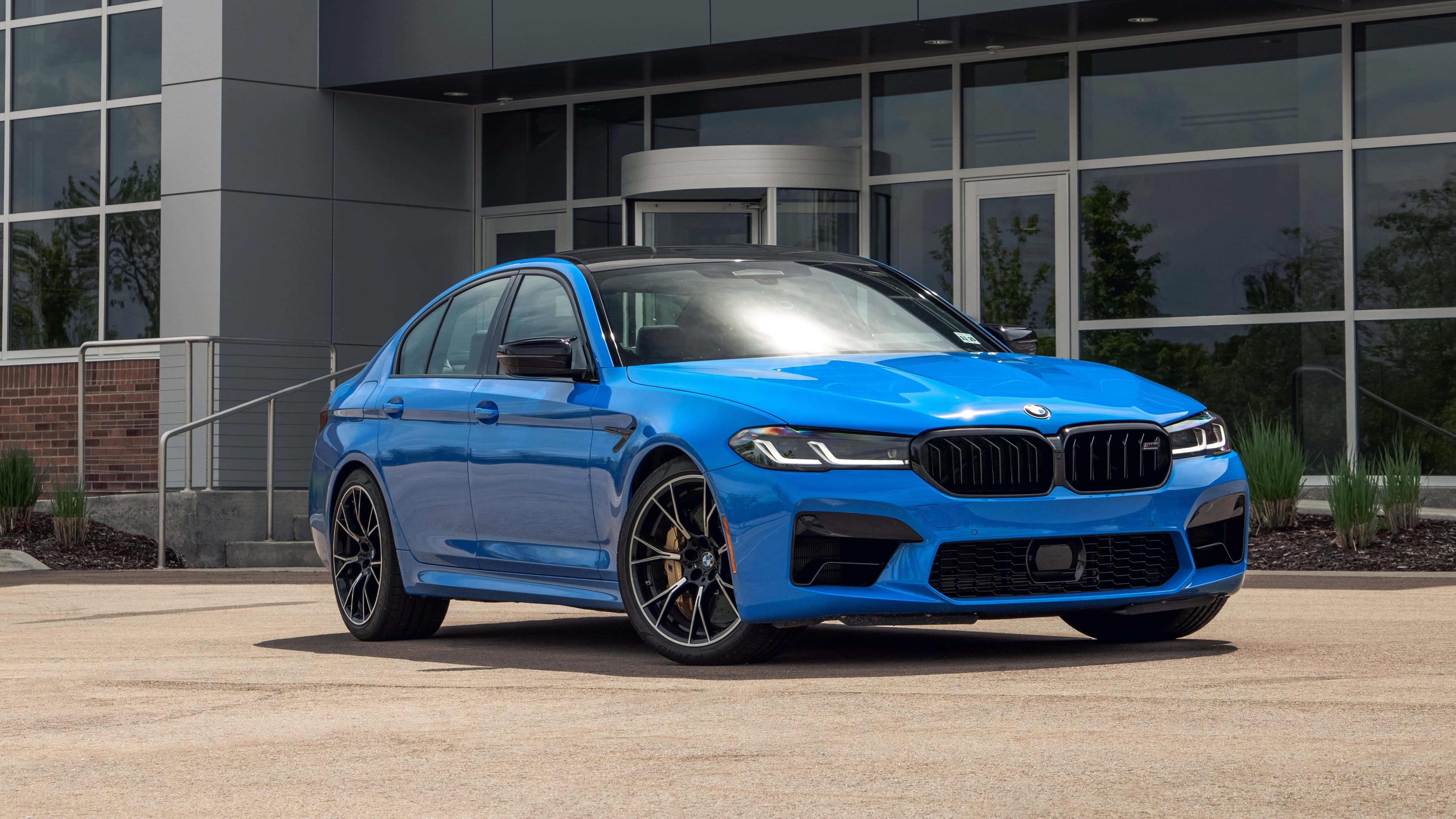 桌布 壁紙 — 一輛充滿活力的藍色BMW M5轎車在日光下閃閃發光，停在一棟現代玻璃建築前。