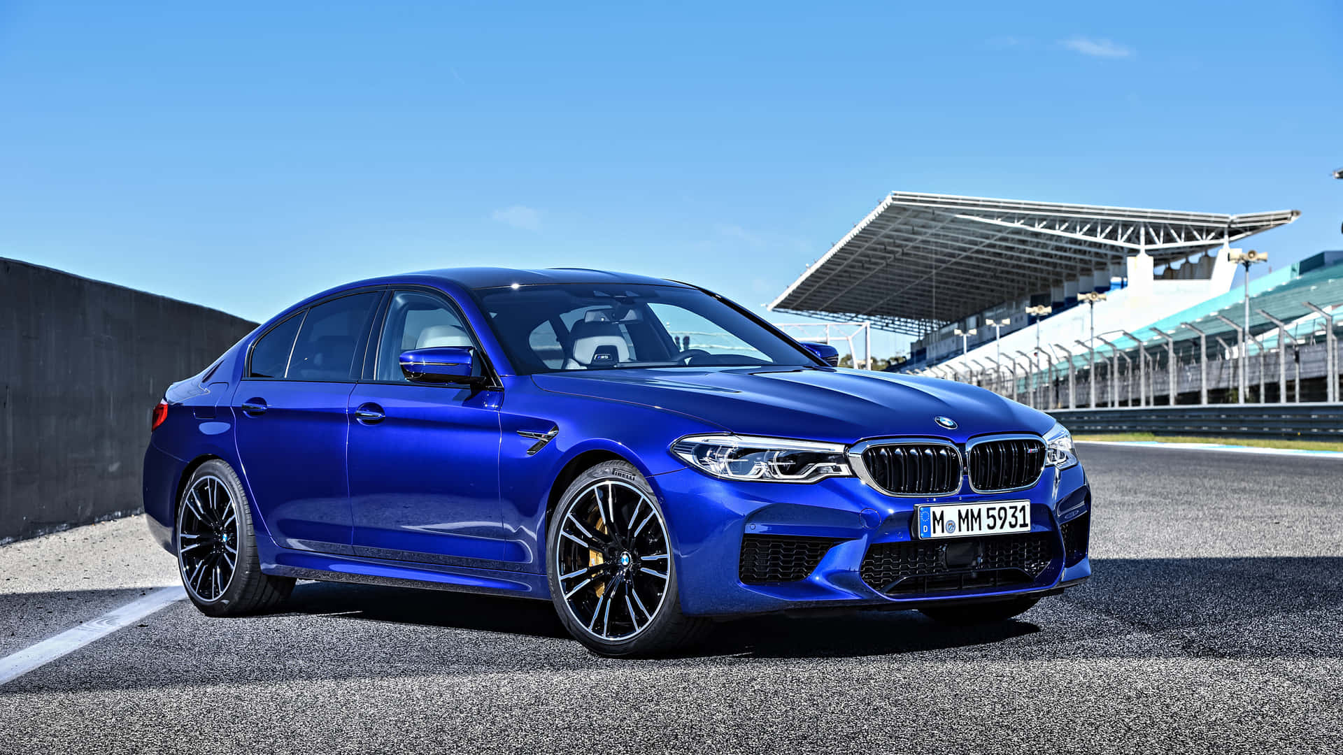 1920x1080 Bmw 桌布 壁紙 — 一輛充滿活力的藍色BMW M5轎車在明亮的陽光下，在瀝青賽道上閃閃發光，背景有看台。