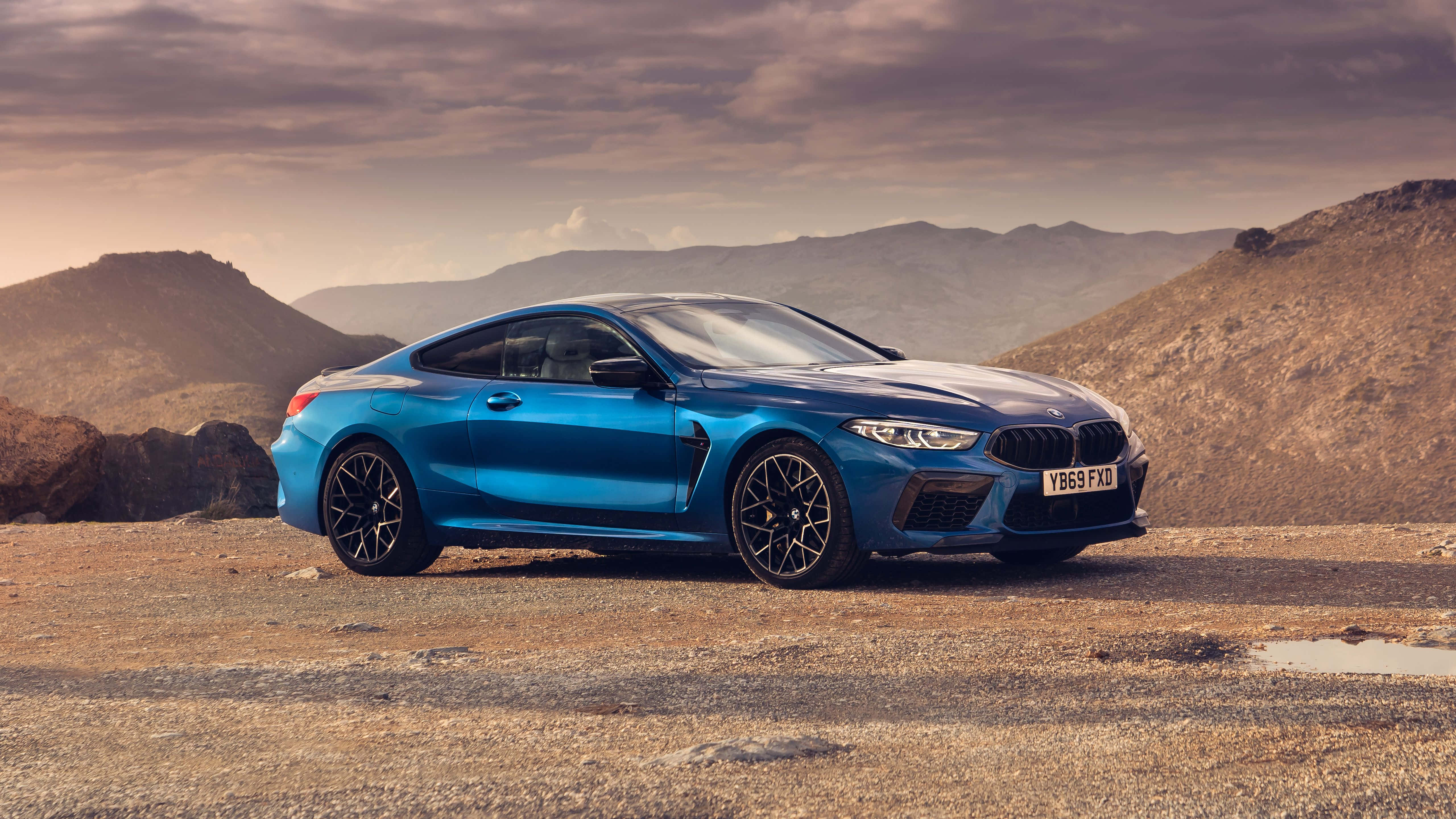 Bmw M8 4K 桌布 壁紙 — 一輛充滿活力的藍色BMW M8 Coupe停放在崎嶇的沙漠道路上，背景是戲劇性的山區地形，天空陰沉。