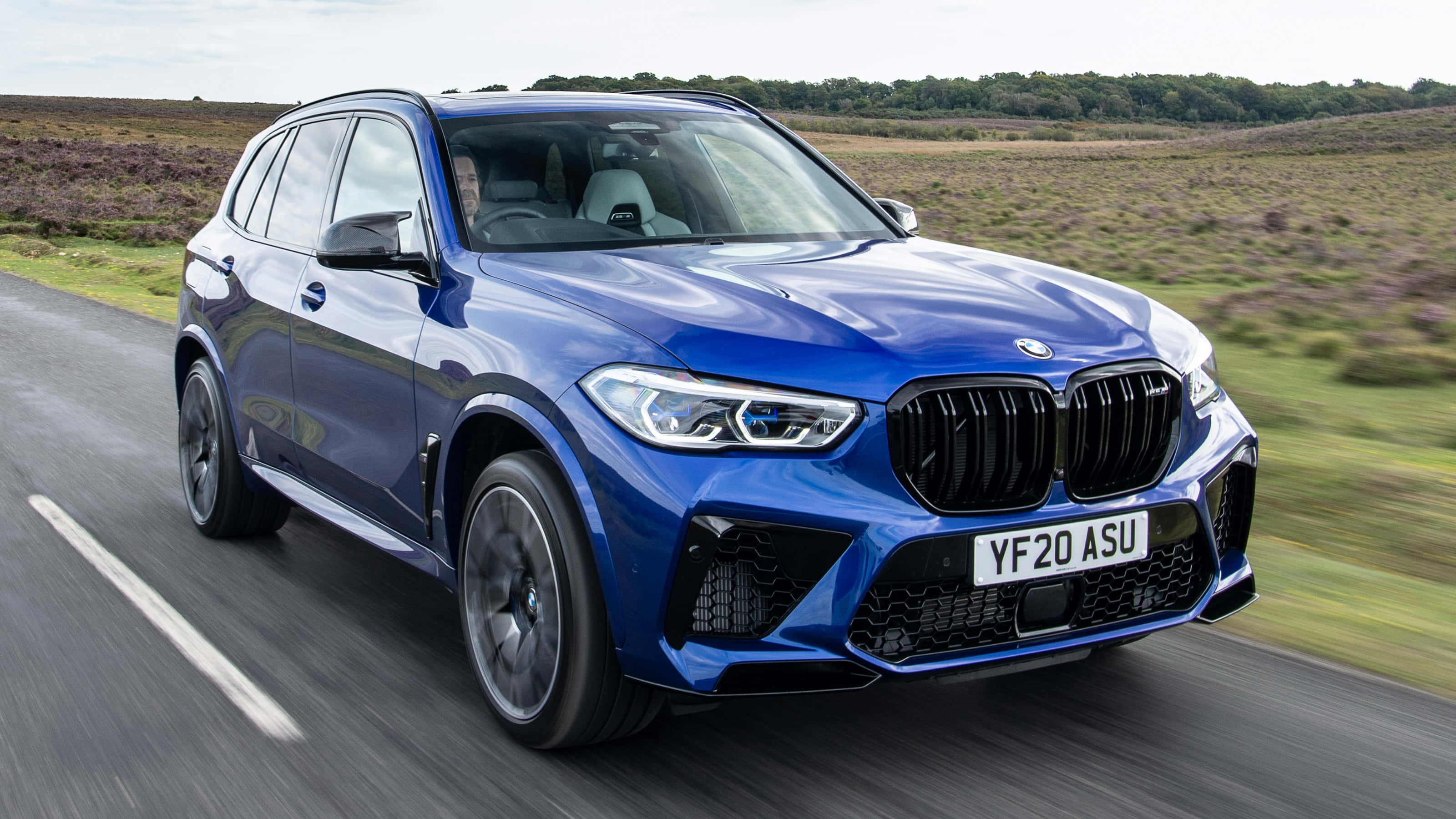 Bmw X5 桌布 壁紙 — 一輛充滿活力的藍色BMW X5 M Competition在蜿蜒的道路上穿過丘陵鄉村景觀飛馳。