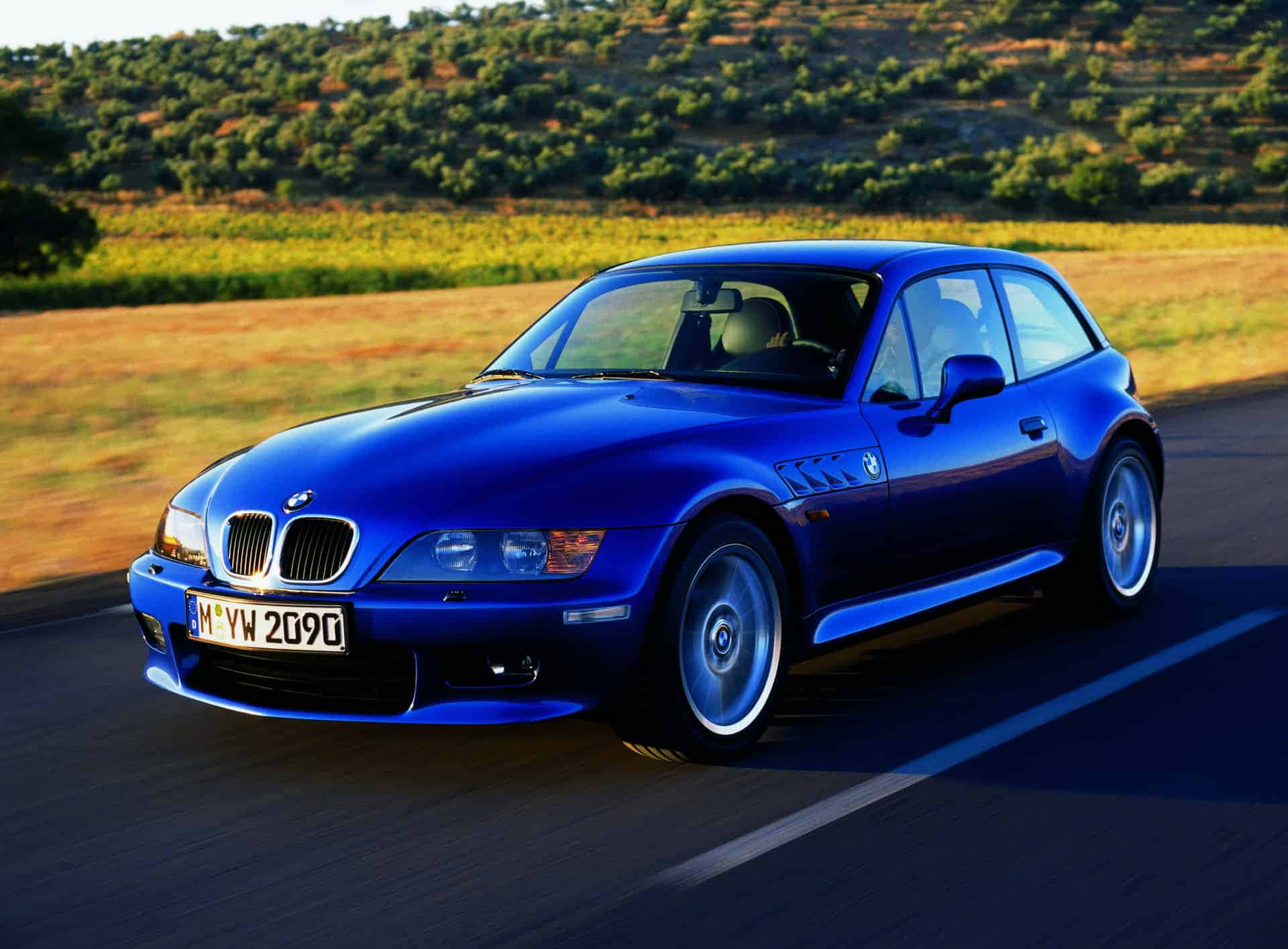 Bmw Z3 桌布 壁紙 — 一輛充滿活力的藍色BMW Z3跑車在陽光明媚的鄉村道路上疾馳，背景是起伏的丘陵和金色的田野。