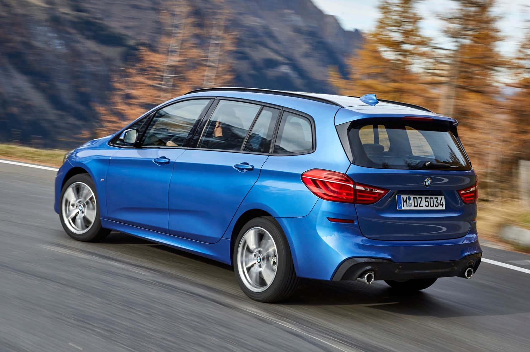 Bmw 2 Series 桌布 壁紙 — 一輛充滿活力的藍色BMW旅行車在秋季樹木和岩石山峰的動態模糊背景中，沿著山路飛馳。