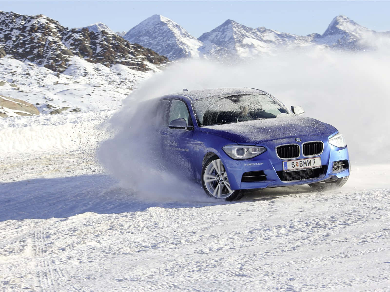 Bmw 1 Series 桌布 壁紙 — 一輛充滿活力的藍色BMW汽車在晴朗的藍天下，在白雪覆蓋的山口疾馳，揚起雪花。