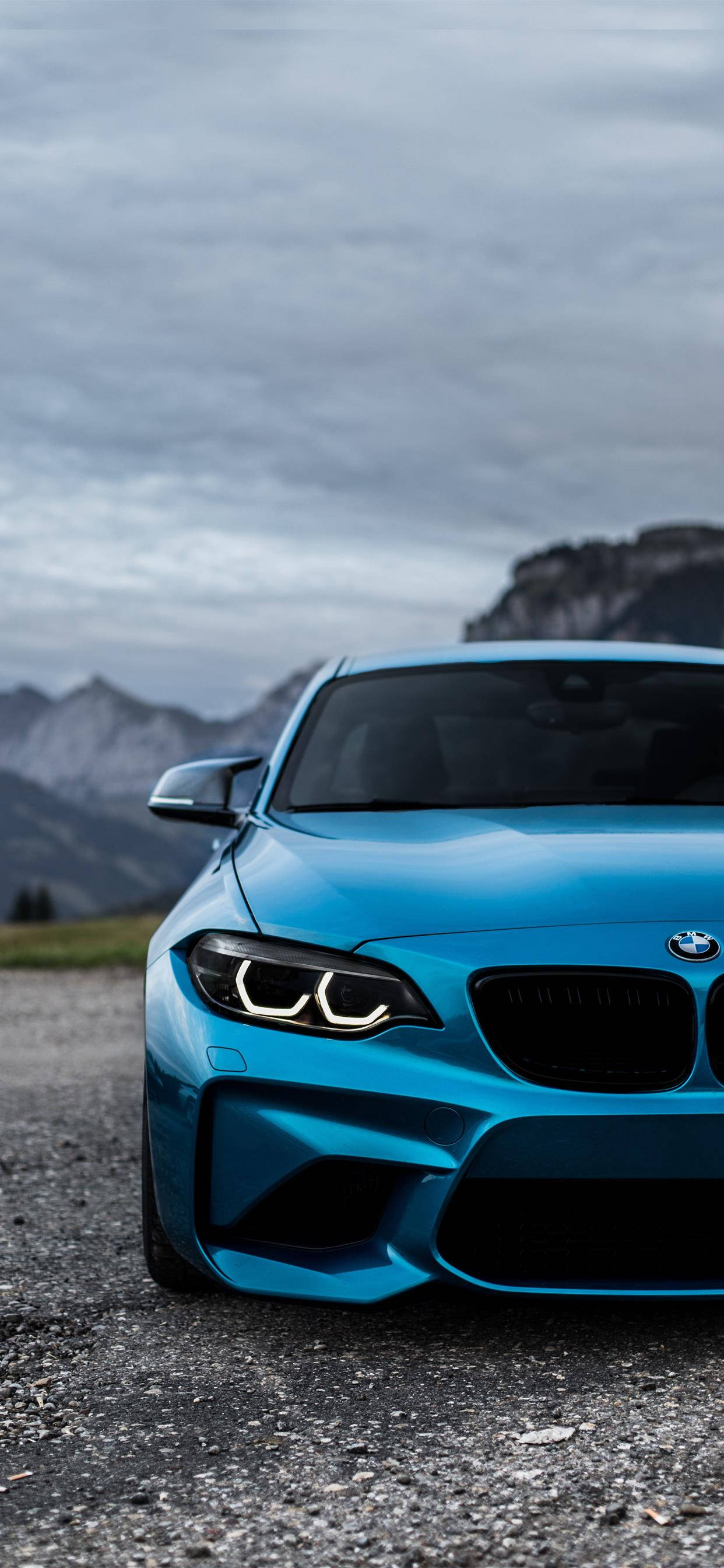 X 桌布 壁紙 — 一輛充滿活力的藍色BMW跑車停在碎石路上，背景是霧氣繚繞的山脈，天空多雲。